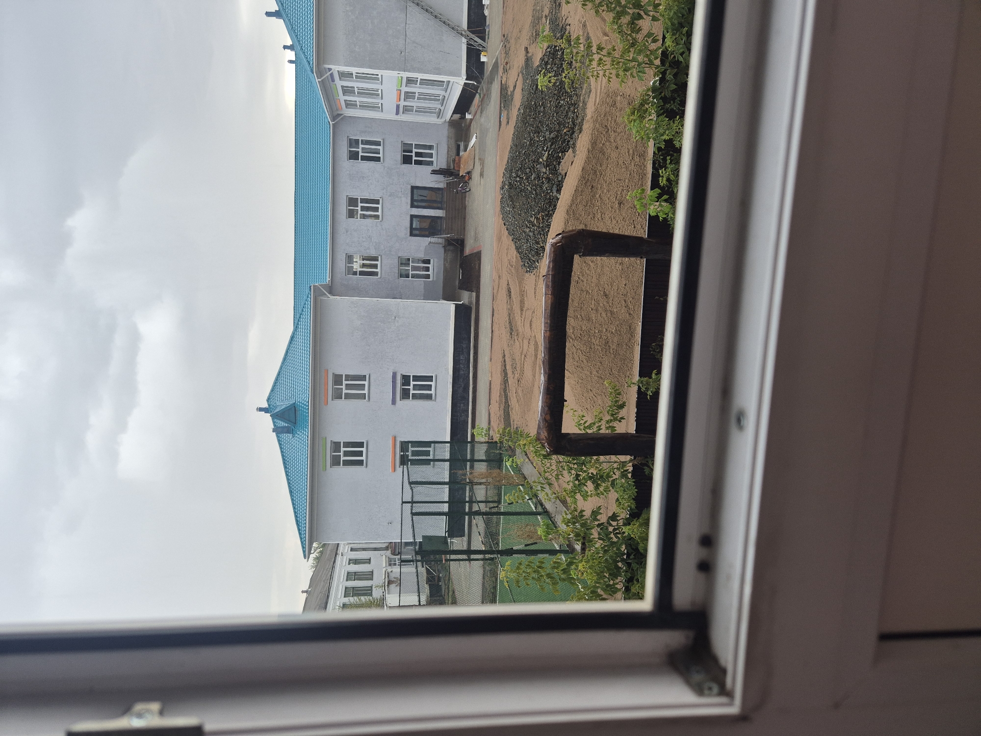The image shows a view from a window overlooking a school building with a bright blue roof. The building is modern and appears to be under construction or recently renovated, with scaffolding visible on the right side. In front of the building, there is a sports field with green turf, surrounded by a fence. Adjacent to the sports field is an area covered in sand and piles of gravel, suggesting ongoing construction or landscaping work. A large metal pipe runs horizontally across the foreground. The sky is overcast. The school is located in Kokshetau, Kazakhstan.