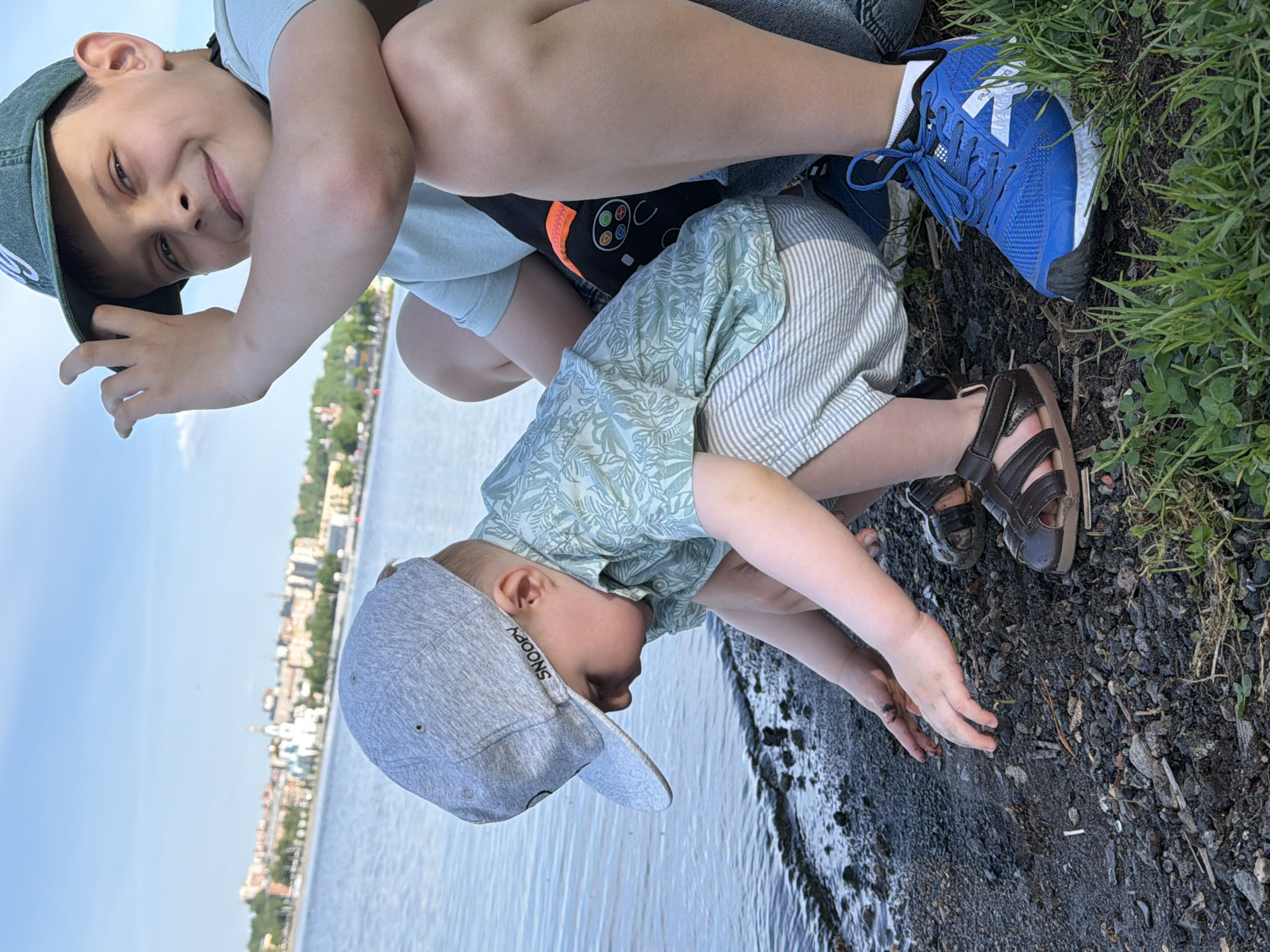 Two children are at the edge of a body of water. One child, wearing a light blue shirt, dark shorts, and blue sneakers, is sitting with legs crossed, partially obscuring the other child. The second child, wearing a light green floral shirt, striped shorts, a grey cap with the word "SNOOPY" on it, and brown sandals, is crouched down near the water's edge. The ground is composed of dark gravel and mud. The background shows the city of Votkinsk, Russia, with buildings and trees visible across the water. The sky is light blue.