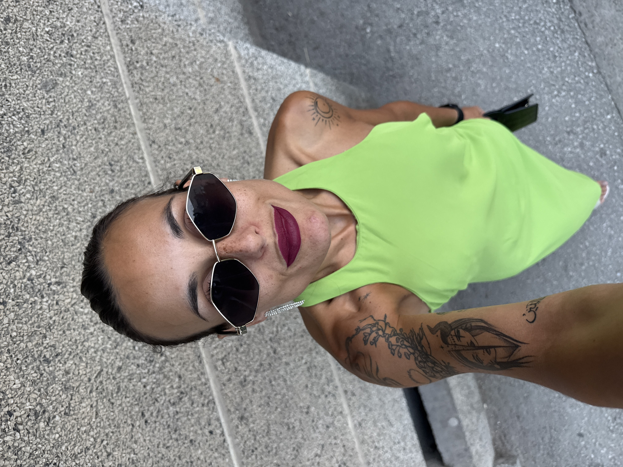 A high-angle, full-body, medium shot captures a woman from the chest up, standing outdoors on a grey, speckled concrete surface. The woman, with her hair pulled back, wears large, polygonal sunglasses with dark lenses and a thin gold frame. She sports dark lipstick and has freckles across her nose and cheeks. A long, dangly, silver earring adorns her right ear. She is wearing a bright lime-green sleeveless dress. Her left arm is visible, displaying a tattoo that wraps around it, featuring a split face design and Arabic script. She also has a smaller tattoo of a crescent moon with radiating lines on her right shoulder. In her right hand, she holds a dark, rectangular object, possibly a phone or wallet. The background consists of textured concrete steps and walls. The scene appears to be located in Pleven, Bulgaria.