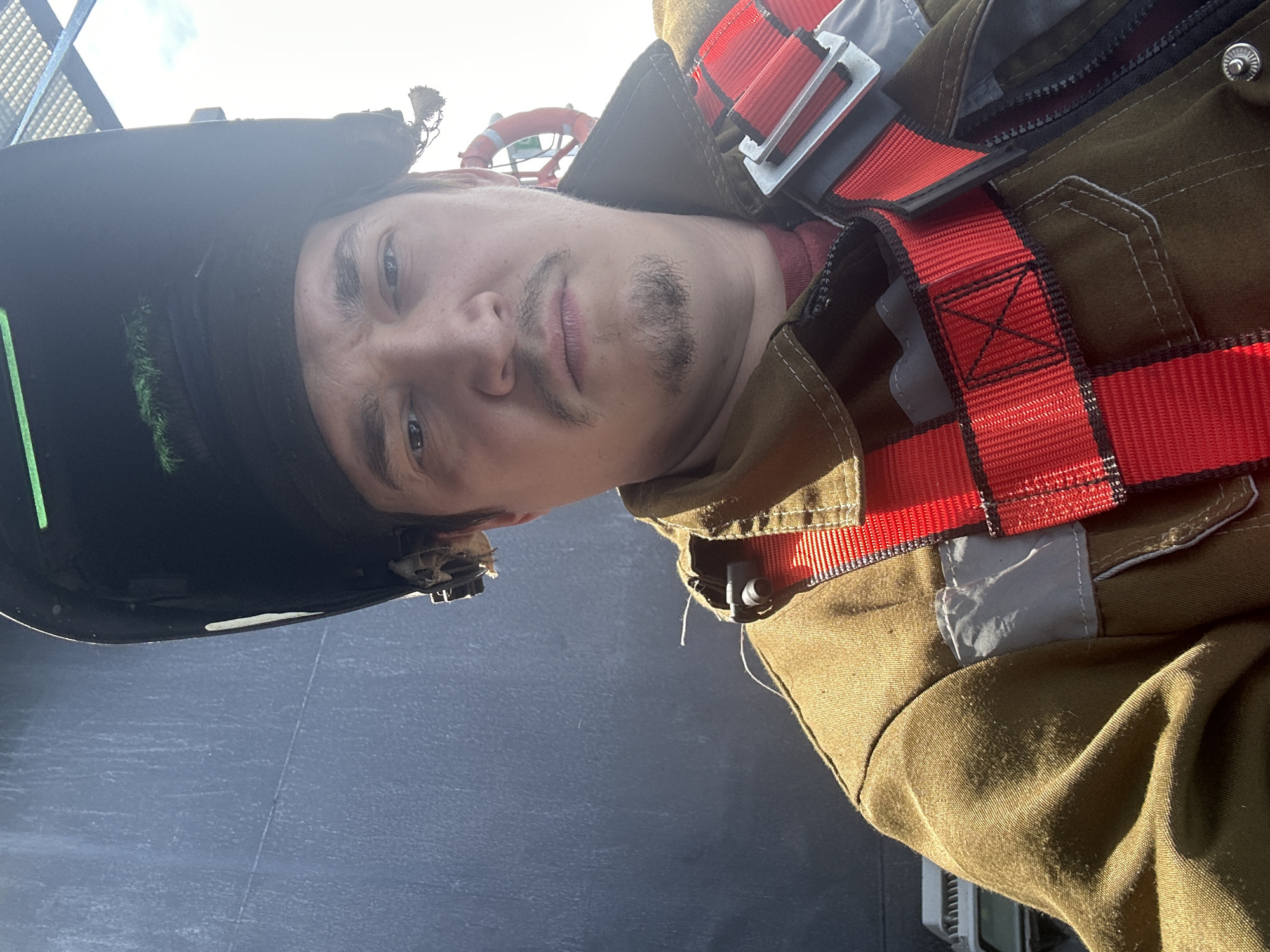 A man wearing a dark brown uniform with red straps, a black helmet with a green stripe, and a thin mustache and beard is pictured from a low angle. He is standing outdoors in daylight against a gray background with a life preserver visible in the upper part of the frame. The location is Unknown, Unknown.