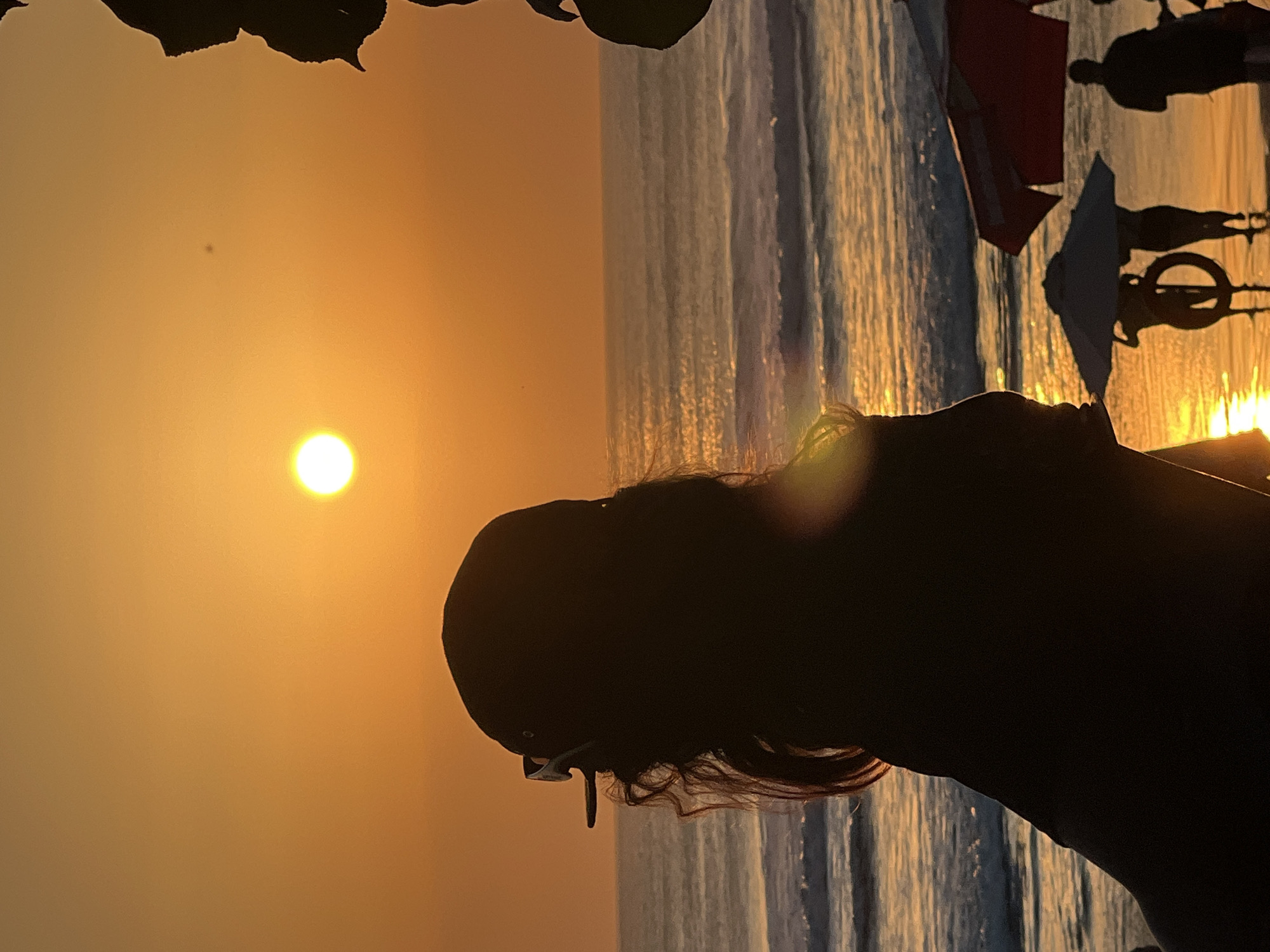 At sunset in Kuta, Bali, Indonesia, a person wearing a baseball cap and sunglasses is silhouetted against the orange sky, facing the ocean. The sun, a bright orb, is visible in the upper part of the frame, reflecting on the water. Waves gently roll onto the sandy beach where other silhouetted figures can be seen under beach umbrellas. The scene evokes a sense of tranquility as the day concludes.