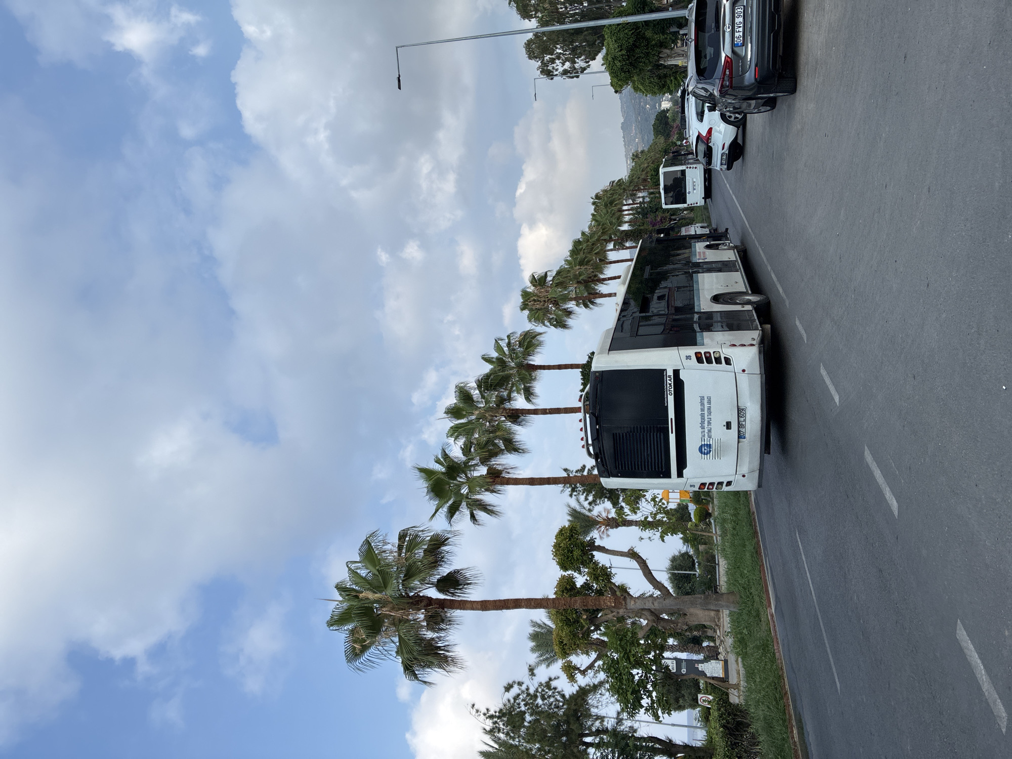 In Alanya, Turkey, on a partly cloudy day, a white bus with the license plate "07 BFL 609" is seen traveling on a road lined with palm trees. The bus has "ALTA BÜYÜKŞEHİR BELEDİYESİ" and "ŞEHİRLİ TOPLU TAŞIMA ARACI" written on its rear. A white car and a dark SUV are also visible on the road, with the SUV displaying the license plate "06 FVG 903". The environment includes a sidewalk, vegetation, and a glimpse of hills in the distance.
