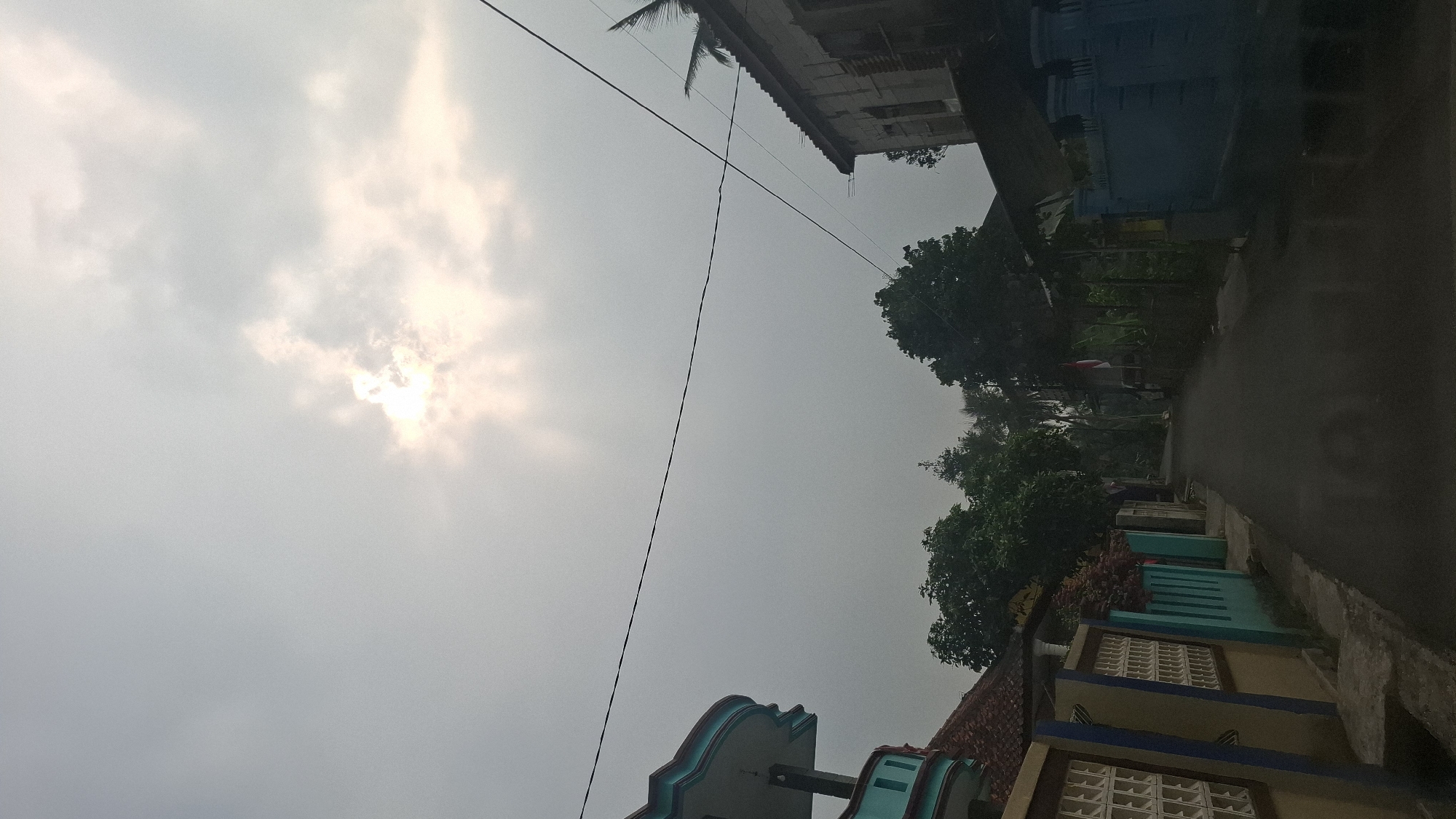 The image depicts a rural street scene in Sukasari, Indonesia, under an overcast sky with the sun partially visible through the clouds. On the left, a building with a distinctive turquoise-trimmed roofline and a beige and blue fence with patterned openings lines the street. To the right, a more weathered building with a corrugated roof and a light blue, solid fence is visible. Dense foliage from trees flanks the sides of the road, which appears to be paved and wet, suggesting recent rain. A single power line stretches horizontally across the upper portion of the frame. The overall atmosphere is subdued, likely due to the cloudy weather. There are no people or visible animals in the frame.