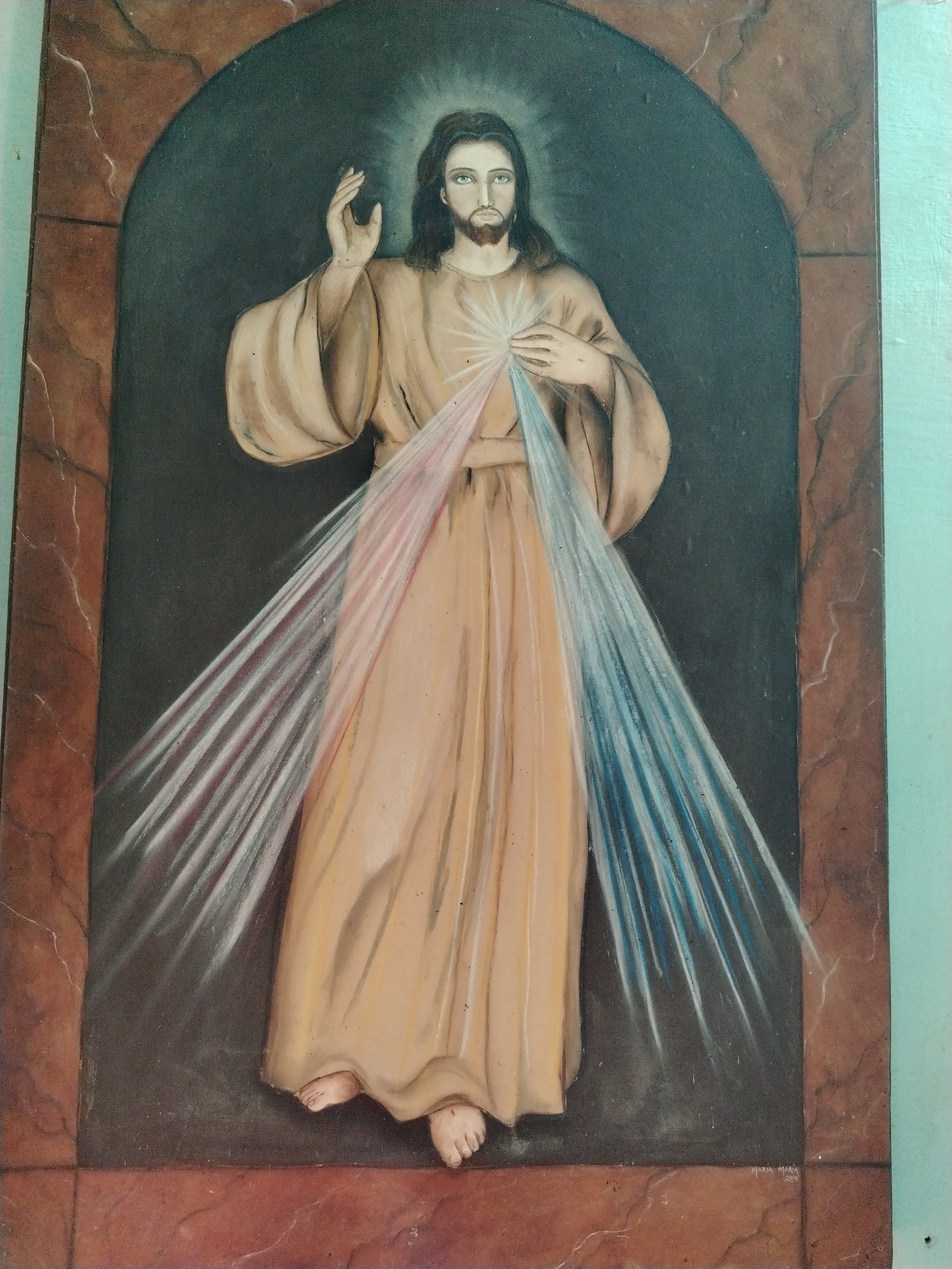 A medium shot shows a painting of the Divine Mercy of Jesus. Jesus is standing in a dark, arched frame. He is wearing a tan robe, with beams of red and blue light emanating from his heart. His right hand is raised in blessing. The painting is framed with brown marble effect. The background is dark. The words "Maria Maria 2010" are visible at the bottom right corner. The painting appears to be hanging indoors. There is no information about the city of Los Leoncitos, Venezuela in the image.
