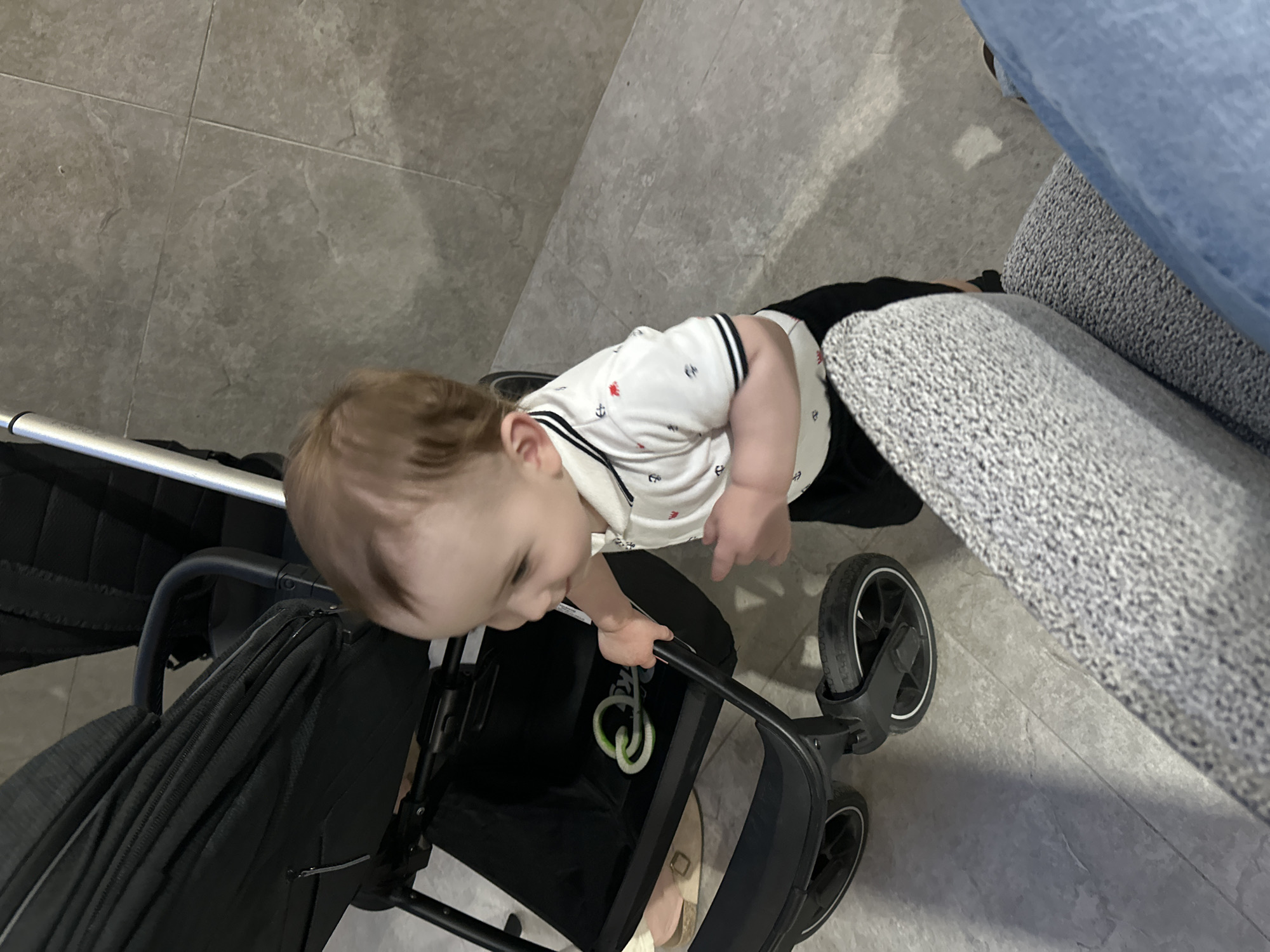 The image shows a toddler standing next to a stroller, holding onto the handle. The toddler is wearing a white polo shirt with a nautical print and black shorts. The stroller is black and has a fabric storage basket underneath. In the storage basket, there is a green, knot-shaped toy. The floor is tiled with large, grey tiles. In the foreground, a person's legs are visible, one wearing blue jeans and the other covered by a textured grey fabric. The background appears to be a wall with a similar grey tiled finish. The setting is likely an indoor space, possibly a home or a public area with a modern interior design. There is no specific information in the image to identify the location as Chișinău, Moldova.