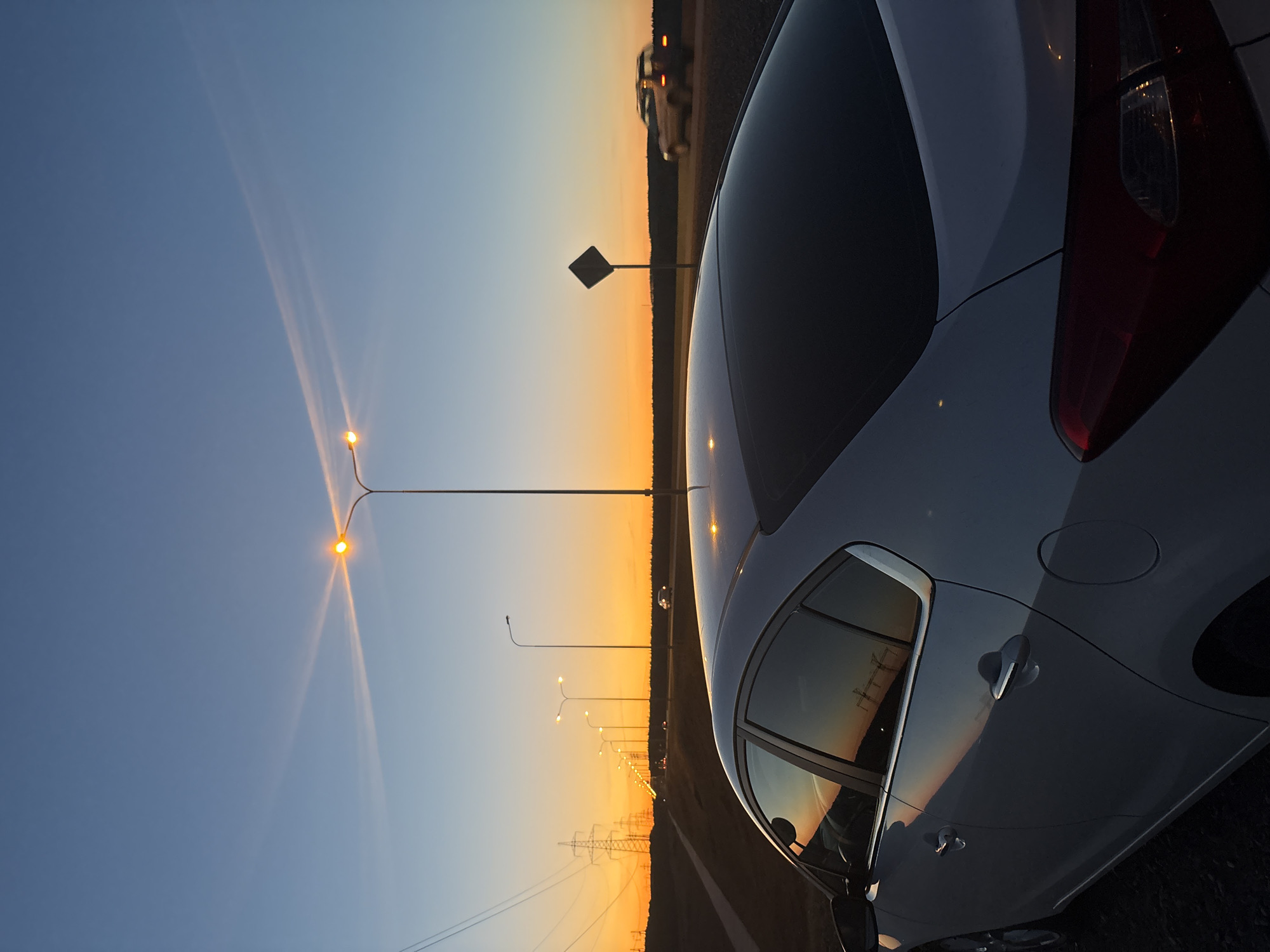 The image presents a partially-framed view of a light-colored car, likely a sedan or hatchback, parked near a highway in Chaikovsky, Russia. The shot is taken at dusk or dawn, as indicated by the gradient of the sky, transitioning from a deep blue to a vibrant orange on the horizon. Several streetlights illuminate the highway, their warm glow contrasting with the cooler tones of the sky. Power lines run parallel to the road. A road sign is visible near the road. Other vehicles can be seen driving along the highway in the distance. The car's taillight is visible. The reflection of the sunset, the powerlines, and other objects are visible on the car's window.