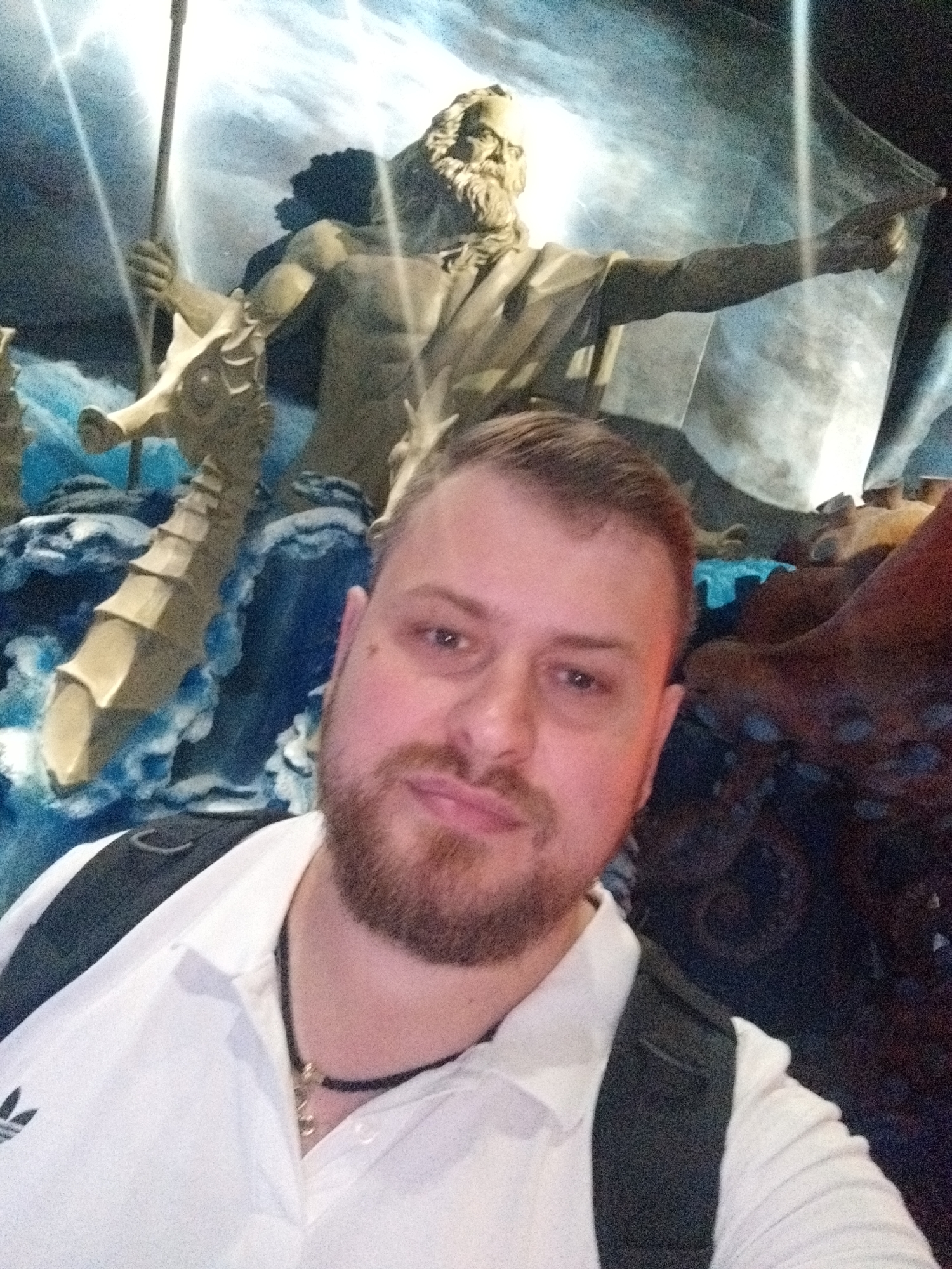 A man is taking a selfie in front of a large statue of Poseidon. Poseidon is depicted with a beard, holding a trident in his left hand and pointing with his right hand. The statue is made of a bronze-colored material and is set against a backdrop of a stormy sky with lightning. The man is wearing a white polo shirt with an Adidas logo and a black backpack. He has short brown hair and a beard. The statue appears to be part of an exhibit, possibly at an aquarium or museum. The specific location in the city of Russky, Russia is not discernible from the image.