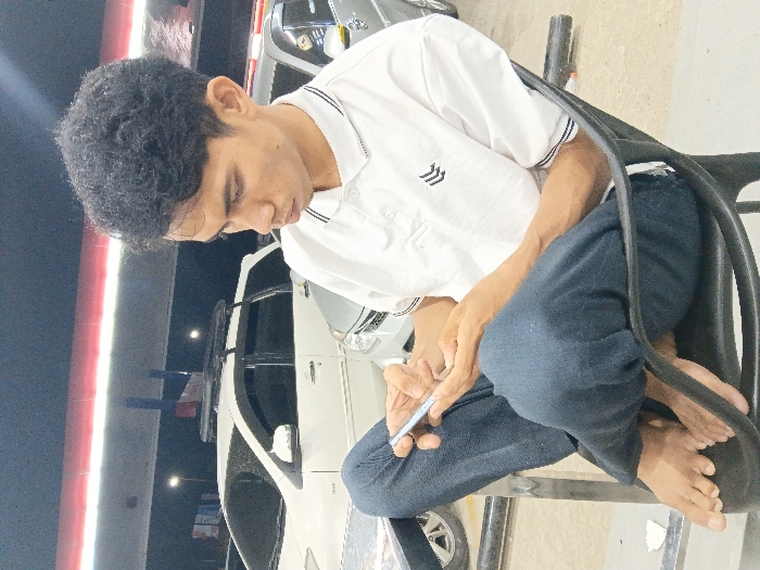 A young adult male is depicted seated barefoot in a black plastic chair, holding a silver smartphone with both hands, and looking down at the device. He is wearing a white short-sleeved polo shirt with a collar featuring black and white stripes, and a black chevron-like logo on the left chest. He has dark blue trousers on, with his right leg crossed over his left. The scene is outdoors at night, under an overhead canopy structure with prominent red and white banding, suggesting a gas station or covered parking area in Seumadam, Indonesia. In the background, a white Toyota Avanza equipped with black roof rails and a silver sedan are visible, parked on a paved surface under the artificial illumination of the overhead lights.