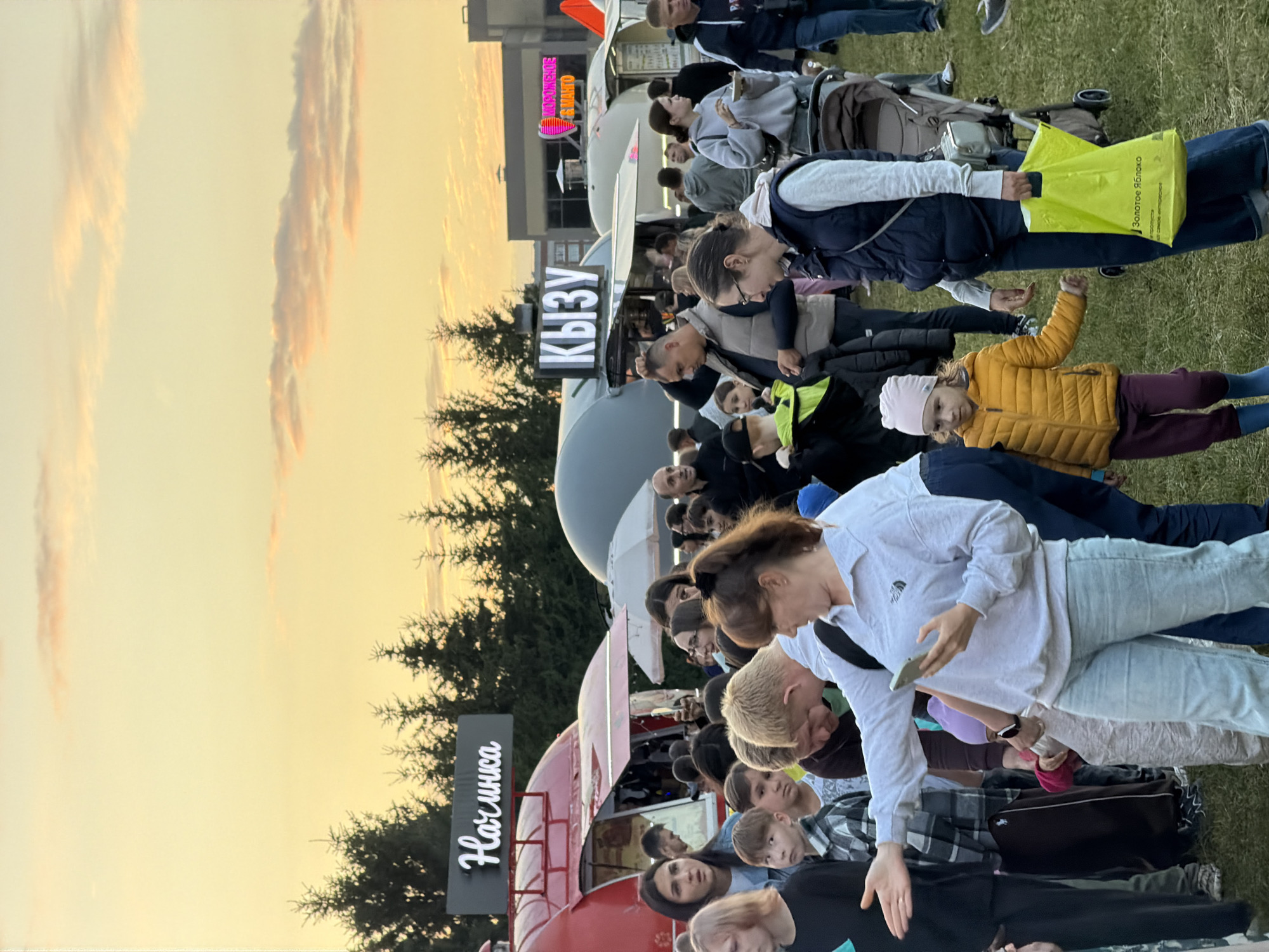An outdoor event is taking place at dusk, with food trucks and a crowd of people. In the foreground, a woman in a light blue sweatshirt and jeans is holding a phone and gesturing with her left hand, while a young girl in a yellow jacket and purple pants stands beside her. To the left, a person in a black shirt and another person wearing a plaid shirt and dark pants are visible. In the background, several food trucks are set up, with illuminated signs above them. One sign reads "Начинка" and another reads "КЫЗУ". Trees line the background, and the sky is a pale orange and yellow, with wispy clouds. Other people are milling around, some looking at their phones, others in conversation. A stroller is also present.