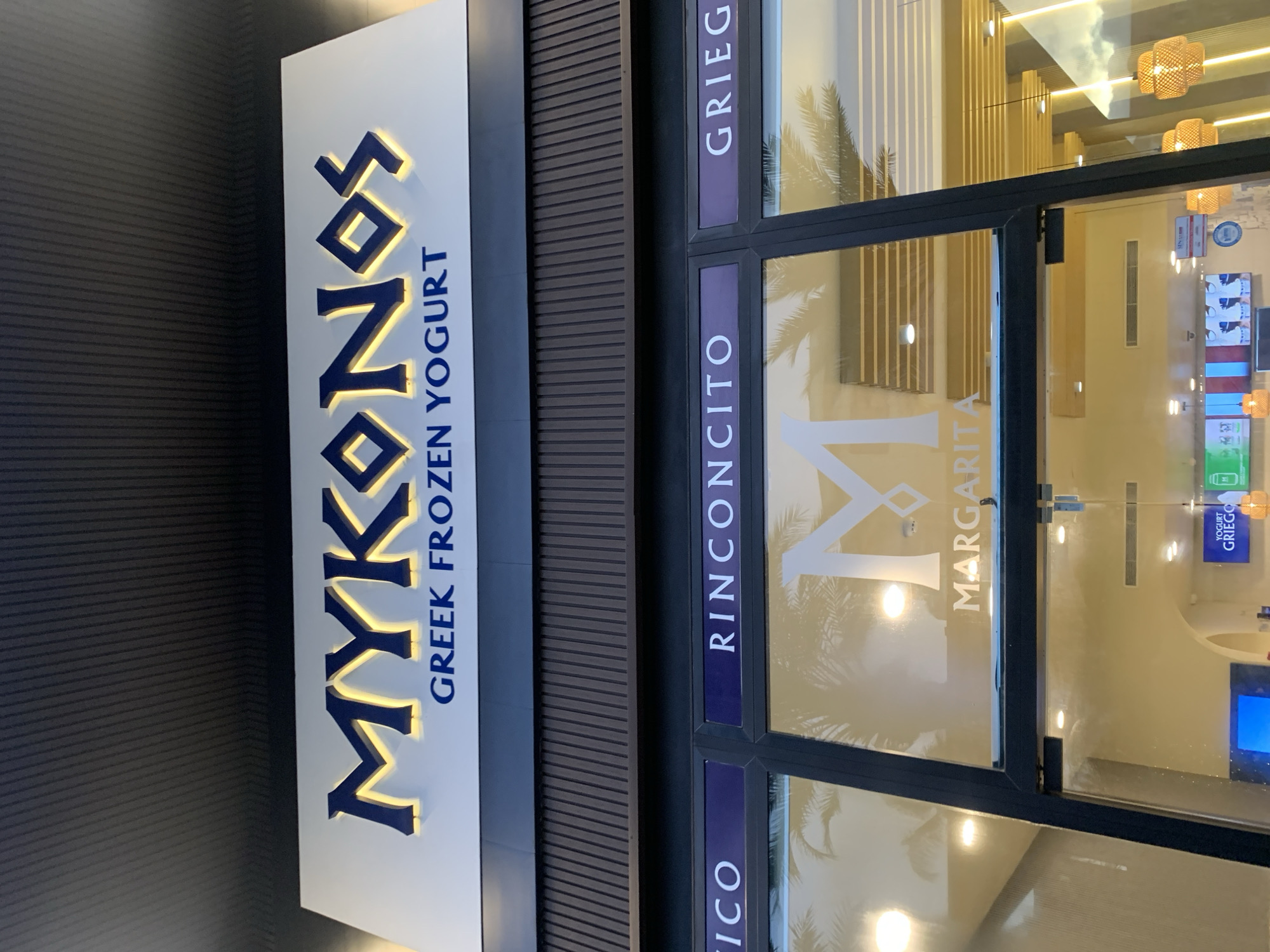 The image displays the brightly lit storefront of a "MYKONOS GREEK FROZEN YOGURT" shop, located in Pampatar, Venezuela. The scene appears to be set at dusk or night, emphasized by the prominent artificial illumination of the sign and the shop's interior.
The main feature is a large, rectangular sign with the shop's name. "MYKONOS" is written in large, stylized blue letters with a warm, yellow internal glow, set against a white background. Below this, the words "GREEK FROZEN YOGURT" are visible in smaller blue text.
Below the sign, the facade features dark-framed glass windows and a dark, ribbed panel. The windows are adorned with dark purple/blue strips containing white text, including "GRIEGO," "RINCÓN CITO," and "MARGARITA," with a partial view of "ICO." A large, stylized white "M" is visible on the main window, reflecting elements like palm trees, suggesting a warm climate.
Through the clear glass, parts of the shop's interior are visible. Warm-toned lighting fixtures, including decorative wicker pendant lights and recessed lights, illuminate the space. Reflections on the windows include a cloudy sky and interior ceiling structures. Inside, several display screens can be seen, one explicitly showing "YOGURT GRIEGO" and others with colorful, illuminated content. No individuals are present in the image, which focuses entirely on the exterior architecture and branding of the modern frozen yogurt establishment.