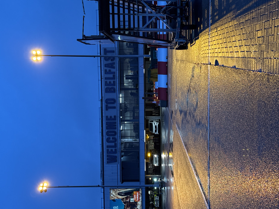 The image depicts the exterior of Belfast International Airport, located in Crumlin, United Kingdom, under twilight conditions. The main airport terminal building is visible, featuring large, illuminated white lettering that reads "WELCOME TO BELFAST" across its upper facade. Below this, the building has a level with multiple glass windows, revealing interior lighting, and a ground-level drive-through or covered parking area. Within this ground level, two white vans are parked, one with amber hazard lights active. A yellow service vehicle is partially visible on the far left. To the left of the building, a large illuminated advertising board displays "easyJet holidays" with an image of an archway.
The scene is lit by two tall metal light poles, each fitted with a cluster of four bright yellow lamps, casting strong illumination onto the foreground. The ground surface consists of wet asphalt with white painted lines and a section of wet, brick-patterned paving on the right. This wetness reflects the artificial lights. In the mid-ground, a series of red and white striped portable barriers are positioned. On the far right, a multi-tiered mobile aircraft boarding staircase or ramp is present, showing its structural components and wheels. No discernible individuals are present in the immediate foreground or mid-ground.
