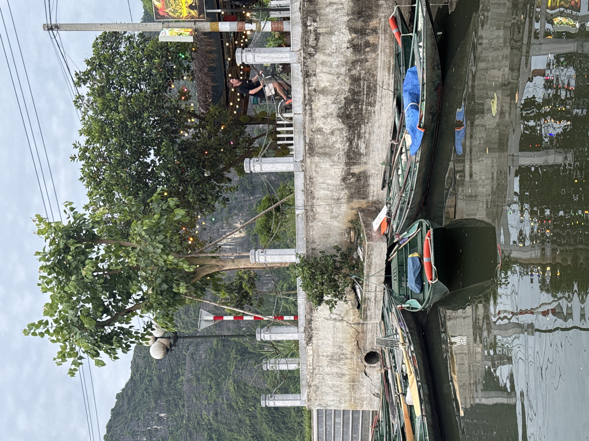 In Nam Hoa Lư Ward, Vietnam, a cluster of green, narrow boats are docked on a calm body of water, their reflections rippling on the surface. A concrete embankment, weathered and cracked in places, rises from the water, topped by a decorative stone railing with pillars and chains. A single tree, adorned with colorful string lights, stands prominently behind the embankment, its branches reaching towards a cloudy sky. A street lamp with three spherical lights is also visible. In the background, a steep, green, mountainous landscape forms a dramatic backdrop. On the embankment, a man is riding a bicycle, his movement captured as he passes by the railing. The scene appears to be a waterfront area, possibly a tourist hub, with elements of natural beauty and human activity.