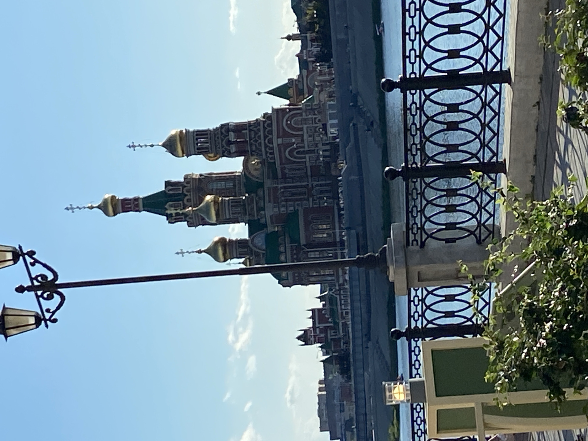 A large, ornate cathedral with multiple golden domes and green spires dominates the background of this outdoor scene. The cathedral is situated on the far bank of a wide river, with a landscaped embankment leading up to it. In the foreground, a black wrought-iron fence with an oval design separates the viewer from the river. A lamppost with two fixtures stands tall to the left of the frame, partially obscuring the cathedral. To the left of the lamppost, a small, square, illuminated lamp is visible. Lush green foliage is present in the lower foreground, casting dappled shadows on the walkway. The sky is a clear, bright blue with a few wispy clouds. The scene appears to be set in Yoshkar-Ola, Russia, and captures a picturesque view of the city's religious architecture.
