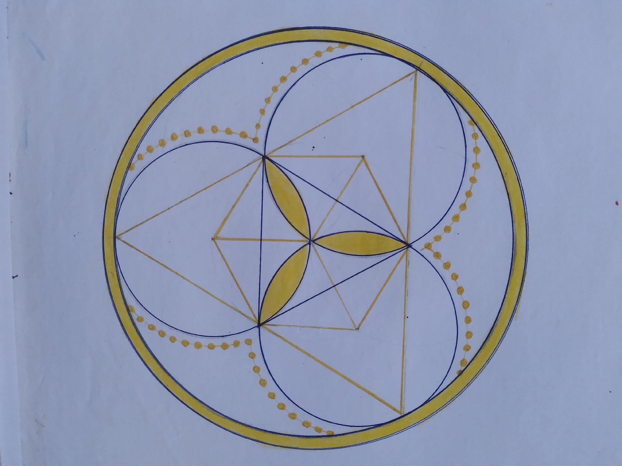 The image displays a hand-drawn geometric pattern on a white surface, possibly paper. The pattern is predominantly composed of yellow and blue lines and yellow colored shapes. The central element is a circle, outlined in yellow with a thinner blue line just inside it. Within this circle, a complex arrangement of triangles and arcs is visible. Three yellow leaf-like shapes are arranged in a radial pattern at the center. Blue arcs connect points on the outer circle, and a string of yellow dots follows these arcs. The drawing appears to be a stylized mandala or sacred geometry design. There is no indication of Baki, Uzbekistan, or any urban environment within the image itself.