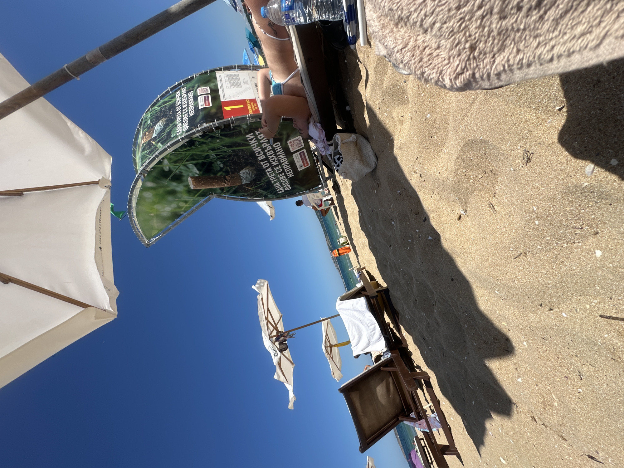 This is a ground-level, tilted shot of a sandy beach scene. The sky is a bright, clear blue, suggesting a sunny day, likely late morning or early afternoon given the strong shadows. The foreground is dominated by sand with scattered seashells. To the left, a white beach umbrella with a wooden frame is partially visible, angled upwards.
In the mid-ground, a circular banner with green foliage and text is displayed. The text, which appears to be in Bulgarian, discusses improper disposal of "fasoves" (likely referring to packaging or similar items) and mentions a "Lifeguard Post." A person is partially visible in the background, wearing a blue swimsuit and lying down. There's also a water bottle and some personal items on a wooden bench or structure behind the banner. Further back, several white beach umbrellas are set up, and beach chairs are visible. The sea and a distant coastline are visible in the far background. The overall mood is relaxed and summery, typical of a beach vacation.