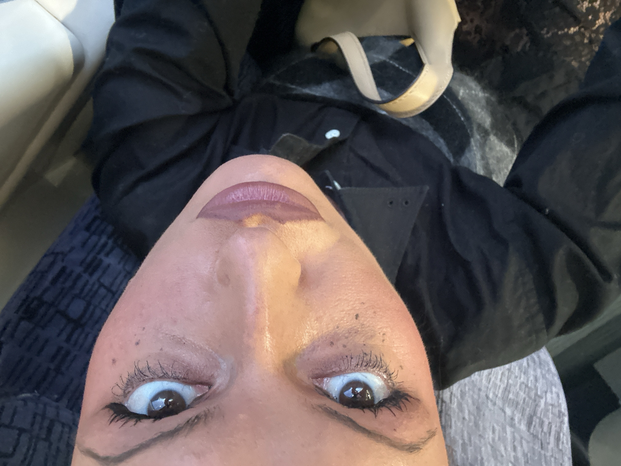The image depicts a close-up, angled selfie of a person's face. The person has fair skin, dark eyes with mascara and eyeliner, and mauve lipstick. There are visible freckles or moles on the face.
The person is wearing a black jacket or coat. In the background, there's a glimpse of a handbag with a tan strap and a checkered fabric. A light-colored surface, possibly a seat, is also visible. The location cannot be determined with only the information provided in the image.