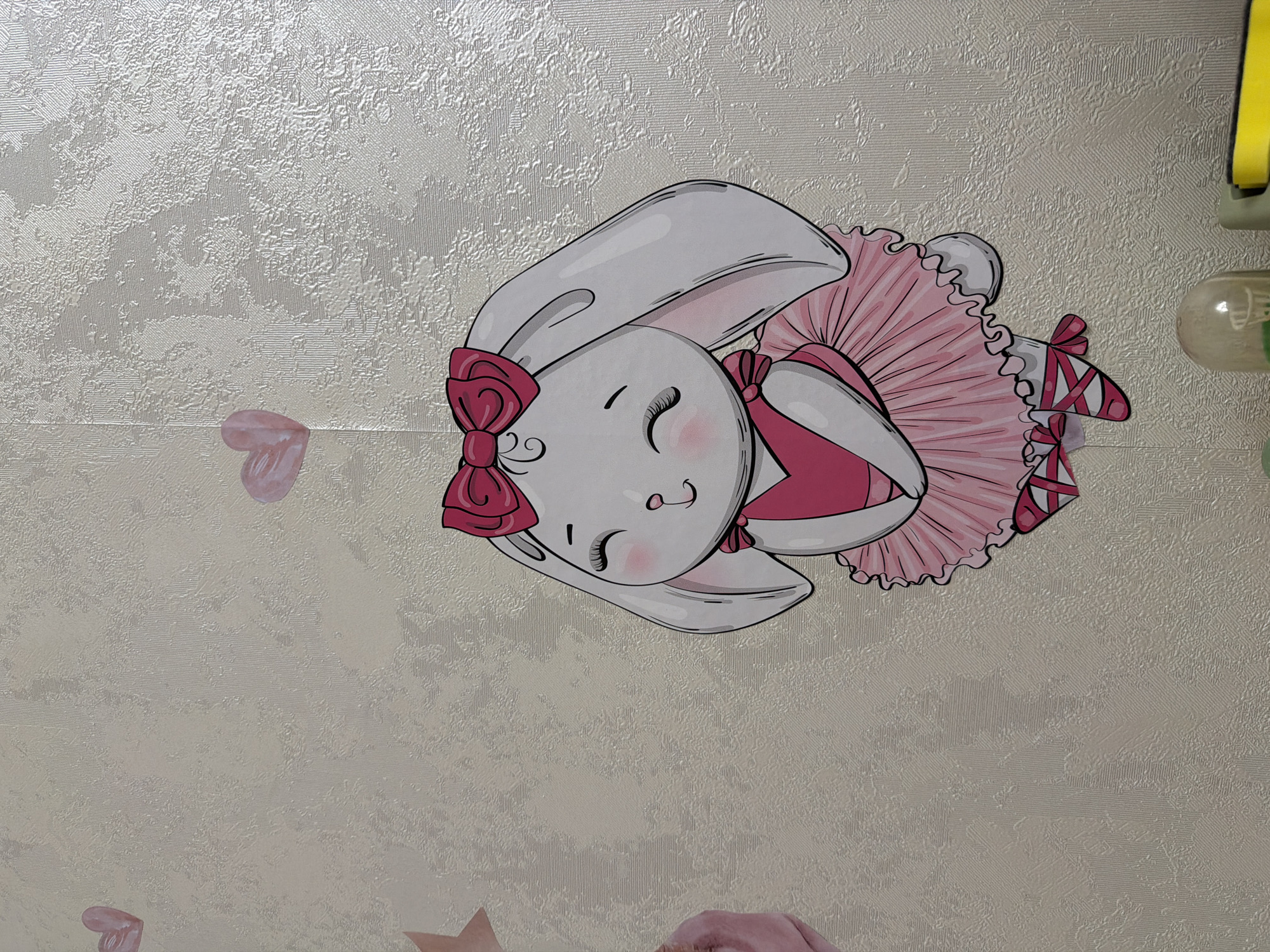 A cartoon drawing of a white rabbit is affixed to a textured, light-colored wall. The rabbit is wearing a pink leotard and a pink tutu. It has a large pink bow on its head and a smaller pink bow at its neck. The rabbit has its eyes closed and a contented expression on its face. Its feet are clad in pink ballet shoes with crisscrossing ribbons. There are also a few pink heart-shaped drawings scattered on the wall. The image appears to be a wall decoration in a child's room or a nursery. The location is Unknown, Unknown.