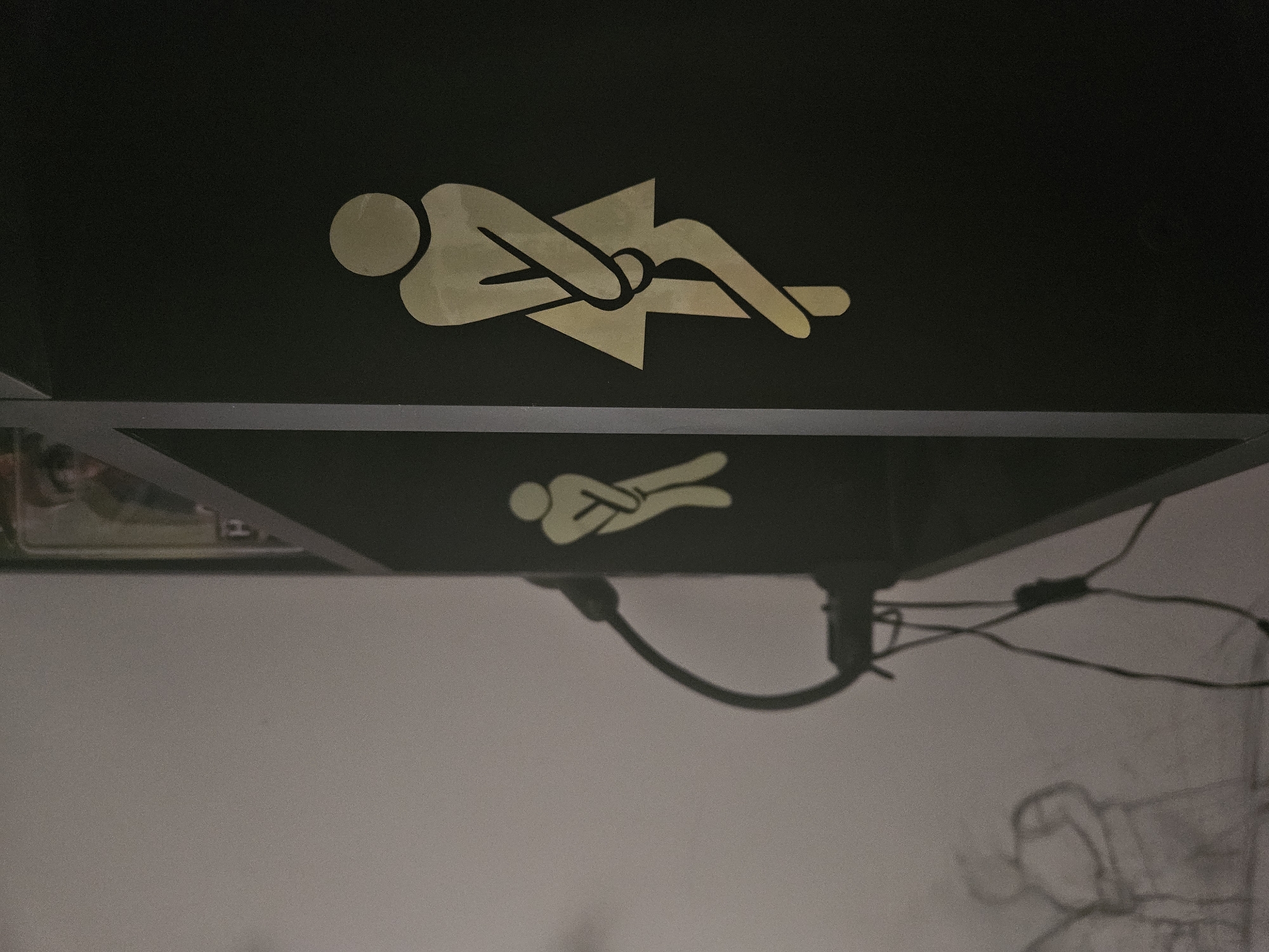 The image shows two figures in silhouette, representing female restrooms. The primary figure on the right is a gold silhouette of a woman with her legs crossed, indicating urgency. This figure is on a black surface, possibly a door or wall. To the left of this figure, a smaller, fainter gold silhouette of a similar figure is visible on a lighter surface. A black cable with a curved attachment runs from the lighter surface towards the black surface, casting a shadow. In the background, on the left side, a faint sketch of figures can be seen on a light-colored wall. The overall scene appears to be an interior location.