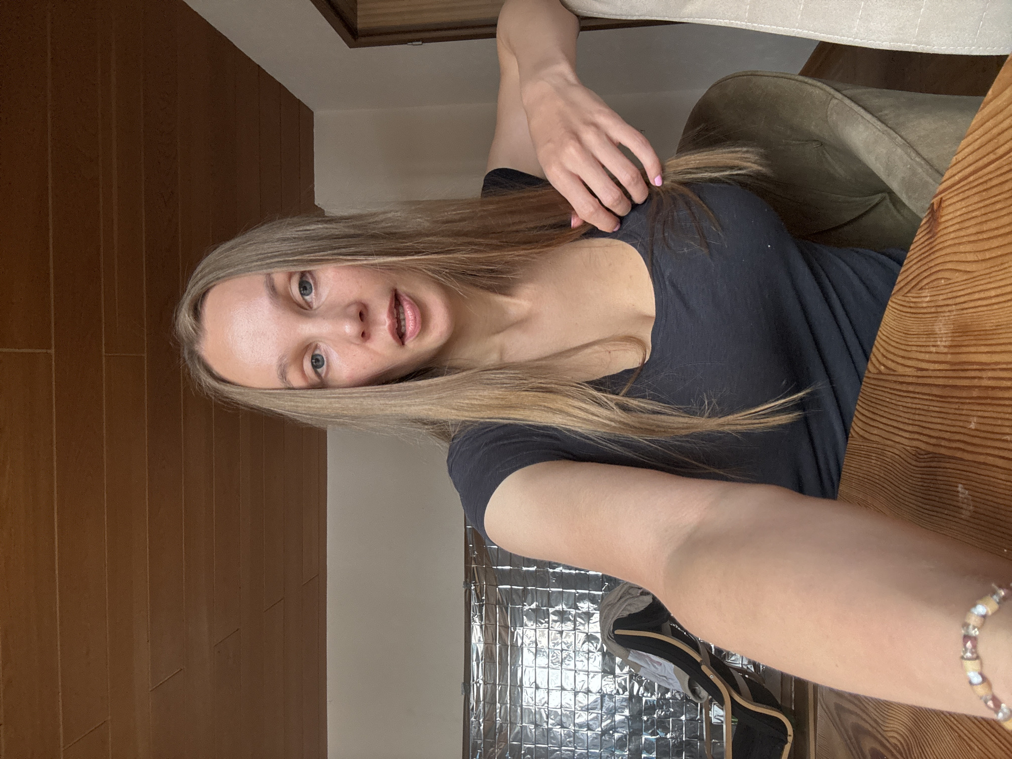 A woman with long blonde hair and blue eyes is seated at a wooden table in a room with wood-paneled walls and ceiling. She is wearing a dark t-shirt and has her left arm extended towards the camera. Her right hand is resting on her chest, playing with her hair. To her left, a metallic-patterned surface is visible on the wall. A green armchair is situated behind her to the right. A light-colored curtain hangs on the far right of the image. The scene appears to be an indoor domestic setting in Überherrn, Germany.