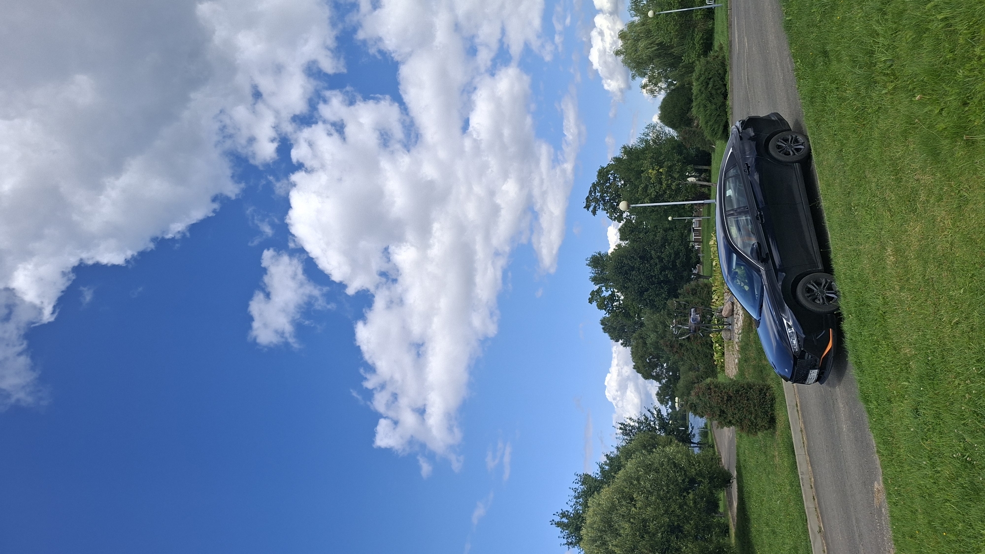 A black sedan is parked on the right side of an asphalt road, angled slightly towards the viewer. The car has orange accents on its front bumper. The road curves to the left in the foreground, bordered by a grassy slope on the right and a manicured lawn on the left. Beyond the lawn, there is a body of water with trees along its bank and further in the background. The background features a line of trees and some structures, including streetlights. The sky above is a vibrant blue with scattered white cumulus clouds. The scene appears to be a park or recreational area in the city of Myadzyel, Belarus, given the presence of water, greenery, and open space.