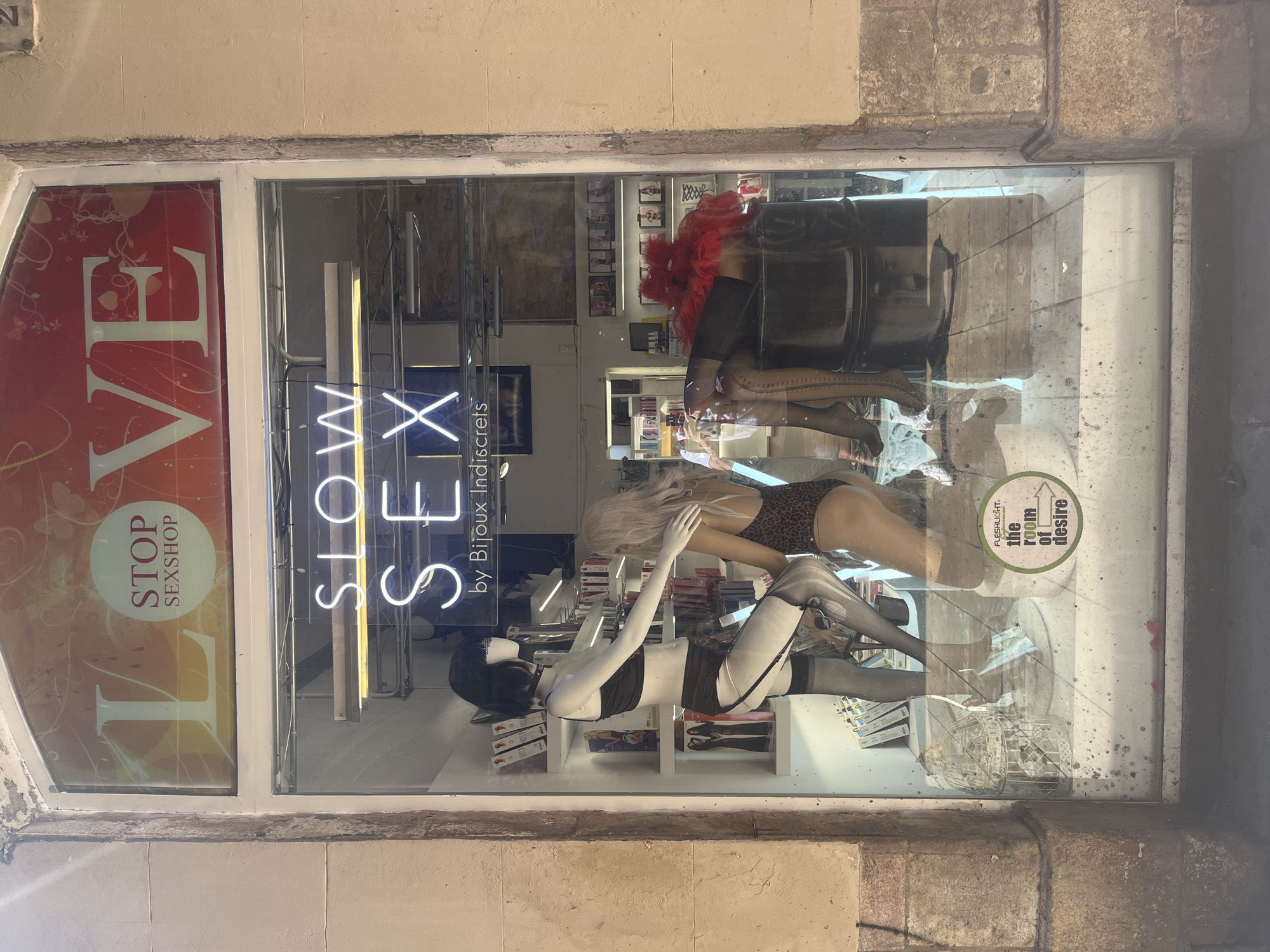 The image depicts the window display of a sex shop in Barcelona, Spain. A window sign with "LOVE STOP SEXSHOP" is visible in the upper left corner of the window, in front of a neon sign reading "SLOW SEX by Bijoux Indiscrets". Inside the display, there are three mannequins dressed in lingerie and hosiery. There is another sign on the glass that reads “Fleshlight, the room of desire.” Additionally, lingerie and other items are visible on shelves in the background.