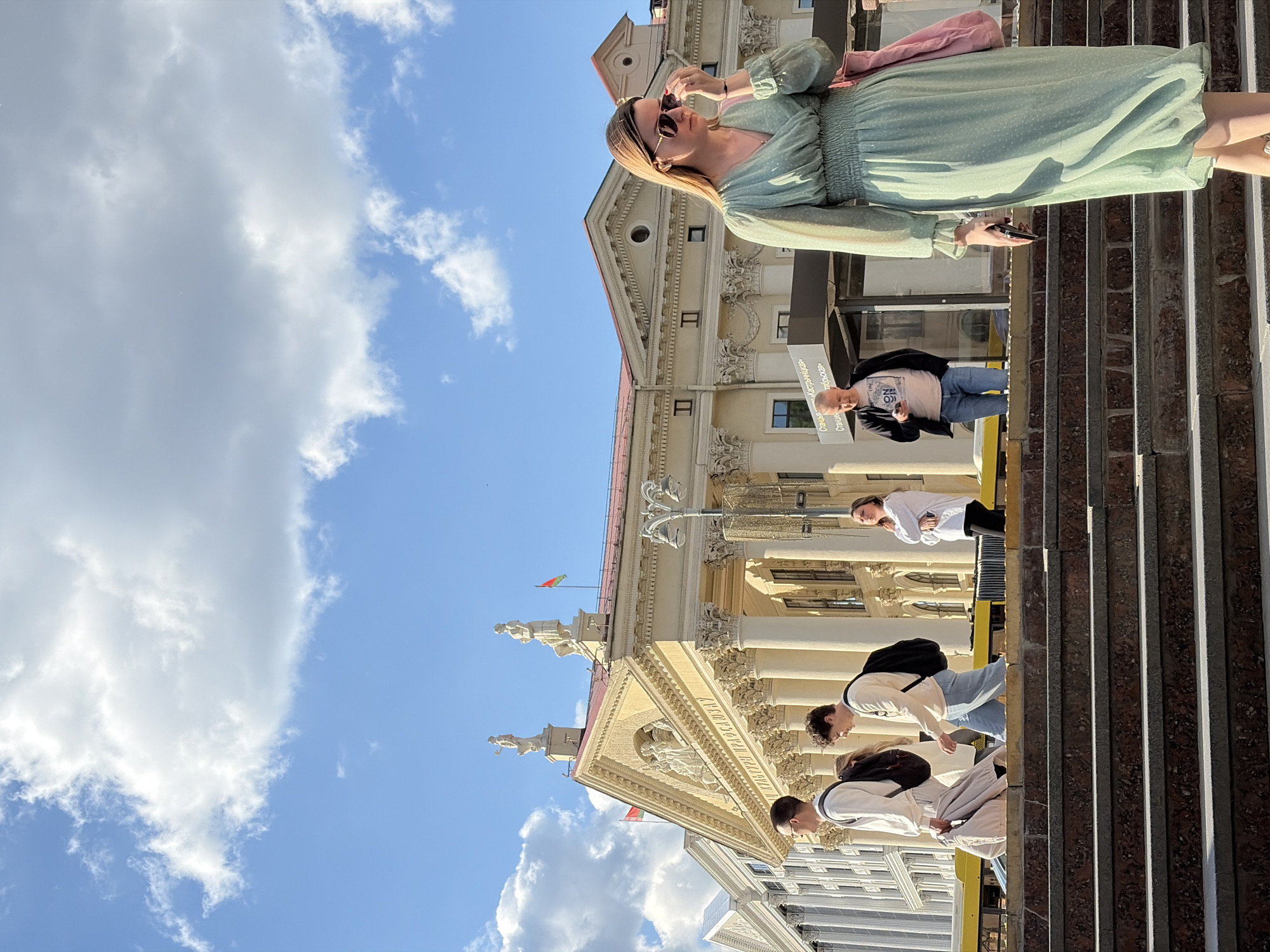 The image shows a view of Minsk, Belarus, with several individuals visible. In the foreground, a woman stands on a set of steps, wearing a light green dress and sunglasses, holding a mobile phone in her right hand and a pink purse on her shoulder. Behind her, a building with ornate architectural details is partially visible. Additional people are walking on the steps, and more architectural elements can be seen, including what appears to be a small flag atop the building. A blue sky with white clouds occupies a significant portion of the upper left quadrant of the image.