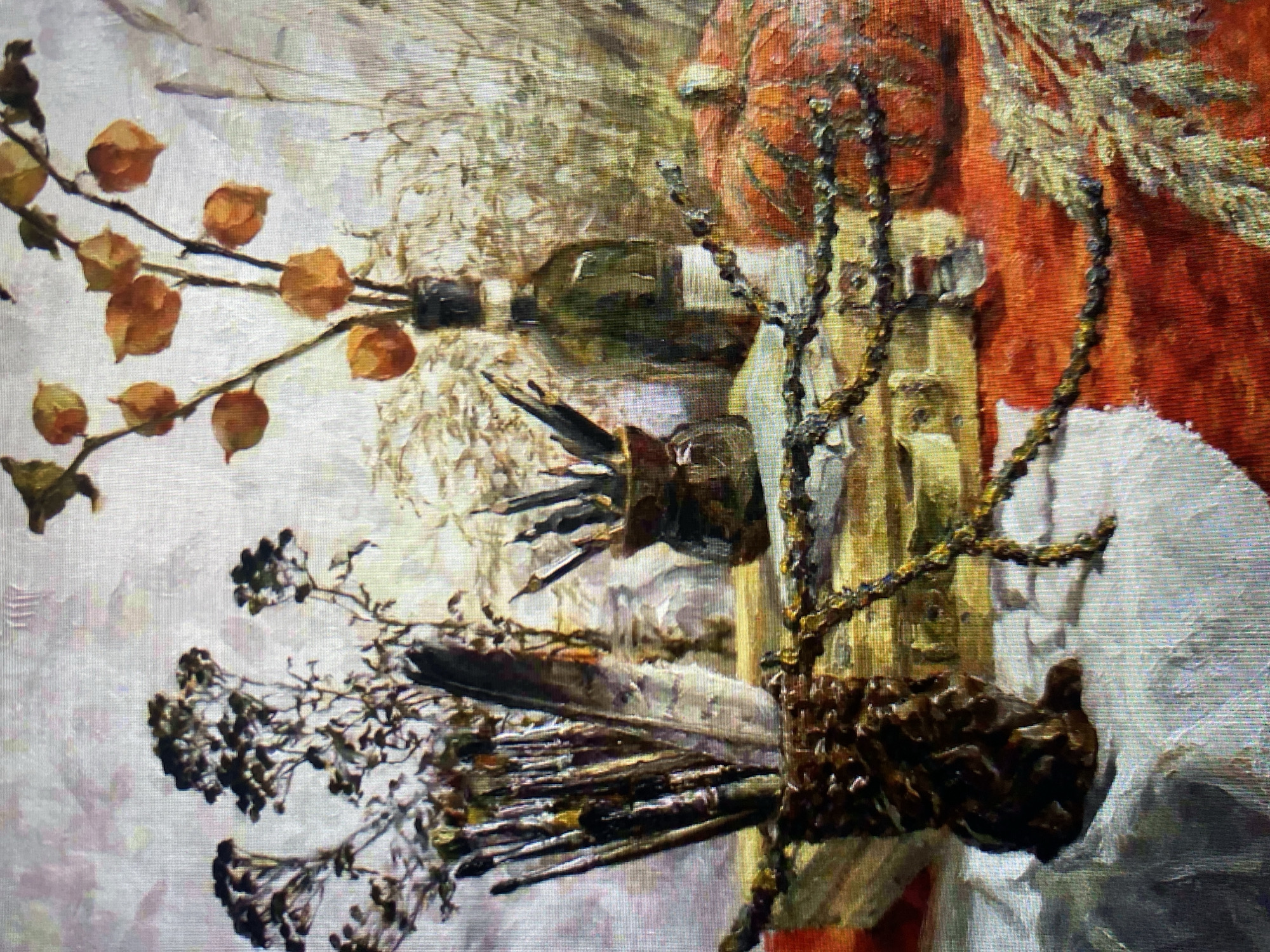 A still life painting features an arrangement of objects on a table, likely in a studio setting, as suggested by the presence of art supplies. The composition includes dried botanical elements, a dark glass bottle, a small pumpkin, and a collection of paintbrushes and pencils. A dark, textured vessel holds the paintbrushes, and another smaller, dark container holds pencils. A single feather rests among the brushes. The objects are placed on what appears to be stacked books, covered by a draped white cloth and an orange fabric. A decorative, chain-like element winds across the foreground, adding a touch of visual interest. The background is textured and suggests a neutral, perhaps painted, wall or backdrop. The lighting creates shadows and highlights, emphasizing the textures and forms of the subjects.