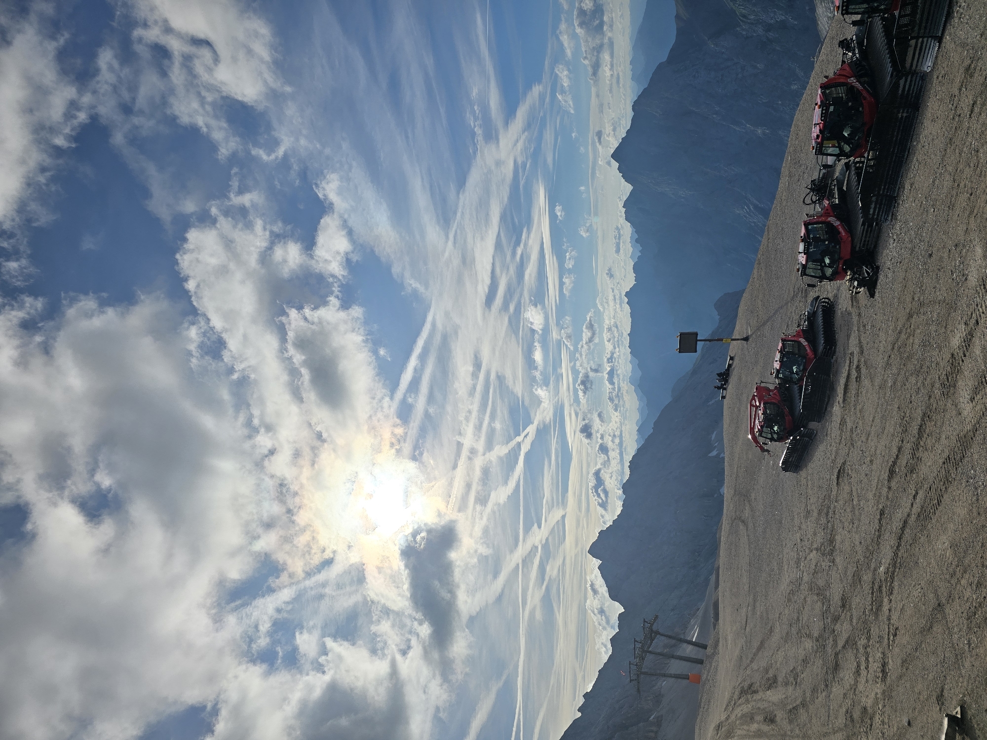 The image depicts a mountain landscape with a clear blue sky filled with scattered clouds and numerous contrails. The sun is partially obscured by clouds, creating a bright halo effect. In the foreground, a gravelly, uneven surface is visible, marked with tire tracks. Several red snow grooming machines are parked on this surface, aligned parallel to each other. In the middle ground, a ski lift structure with towers and cables stands on the left. A warning sign on a yellow post is situated further down the slope. The background features a range of imposing, dark blue mountain peaks. The scene is likely the Zugspitze plateau near Garmisch-Partenkirchen, Germany, given the presence of snow groomers and ski lift infrastructure in a high alpine environment. There are a few silhouetted figures visible in the distance, sitting near the warning sign and on a bench, but no significant interactions or actions are depicted. The overall environment suggests a high-altitude, mountainous area, possibly during the off-season for snow sports or in preparation for it.