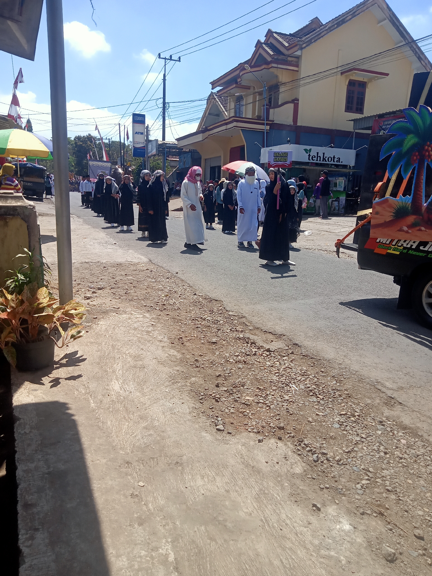 This image depicts a procession of people walking on a street in Pule, Indonesia. The individuals are predominantly dressed in traditional Islamic attire, with many wearing black robes and head coverings. Some are also wearing white robes and headscarves, suggesting a mix of participants or different roles within the procession.
The procession is moving along a paved road, with a dirt and gravel shoulder on the left side of the image. In the background, there are buildings, including a light yellow building with a dark blue and red trim, and a sign that reads "tehkota.". There are also utility poles and overhead wires visible, indicating an urban or semi-urban setting.
On the right side of the image, a portion of a vehicle, possibly a truck, is visible. It has an illustration of a palm tree and text that reads "MITRA JIHA".
The overall scene appears to be a public event or religious gathering taking place during the daytime, under a clear blue sky with some clouds. The lighting suggests it is sunny. The presence of a BRI ATM sign on a pole indicates a commercial area.