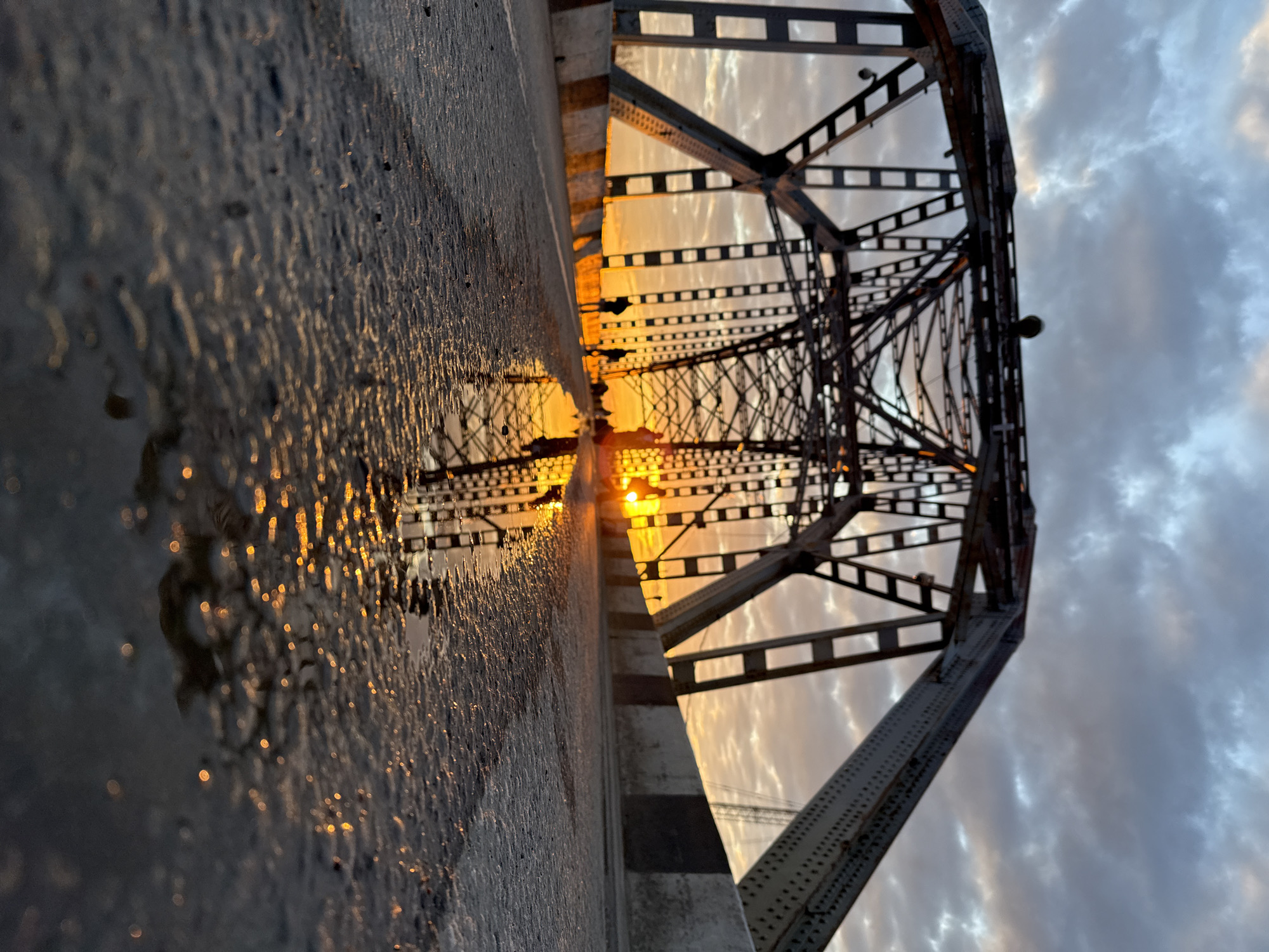 The image captures a dramatic, low-angle perspective of an old metal truss bridge in Dmitrov, Russia, likely during sunrise or sunset. The sun, a brilliant orange and yellow orb, is partially obscured by the bridge's intricate lattice work, casting a warm glow across the scene.
The foreground features a wet, dark, textured surface, presumably the bridge deck or an adjacent pathway, which reflects the sun's light in numerous sparkling bokeh-like glints. These reflections create a shimmering path leading towards the bright light source. The metallic structure of the bridge, with its riveted beams and crossbars, dominates the central and right portions of the frame, silhouetted against the bright sky and illuminated by the low sun.
The sky, particularly on the right side and visible through the bridge's framework, is filled with soft, scattered clouds, painted with hues of gray, white, and a subtle orange from the sun's diffused light. The overall atmosphere suggests a tranquil moment, highlighting the interplay between the industrial architecture and natural light.
No people or explicit activities are visible, emphasizing the structural grandeur of the bridge and the natural phenomenon of the sun's position. There is no discernible text present in the image. The wet surface suggests recent rain or heavy dew, adding to the reflective quality of the scene. The image is composed with a strong diagonal tilt, giving it a dynamic and immersive feel.
