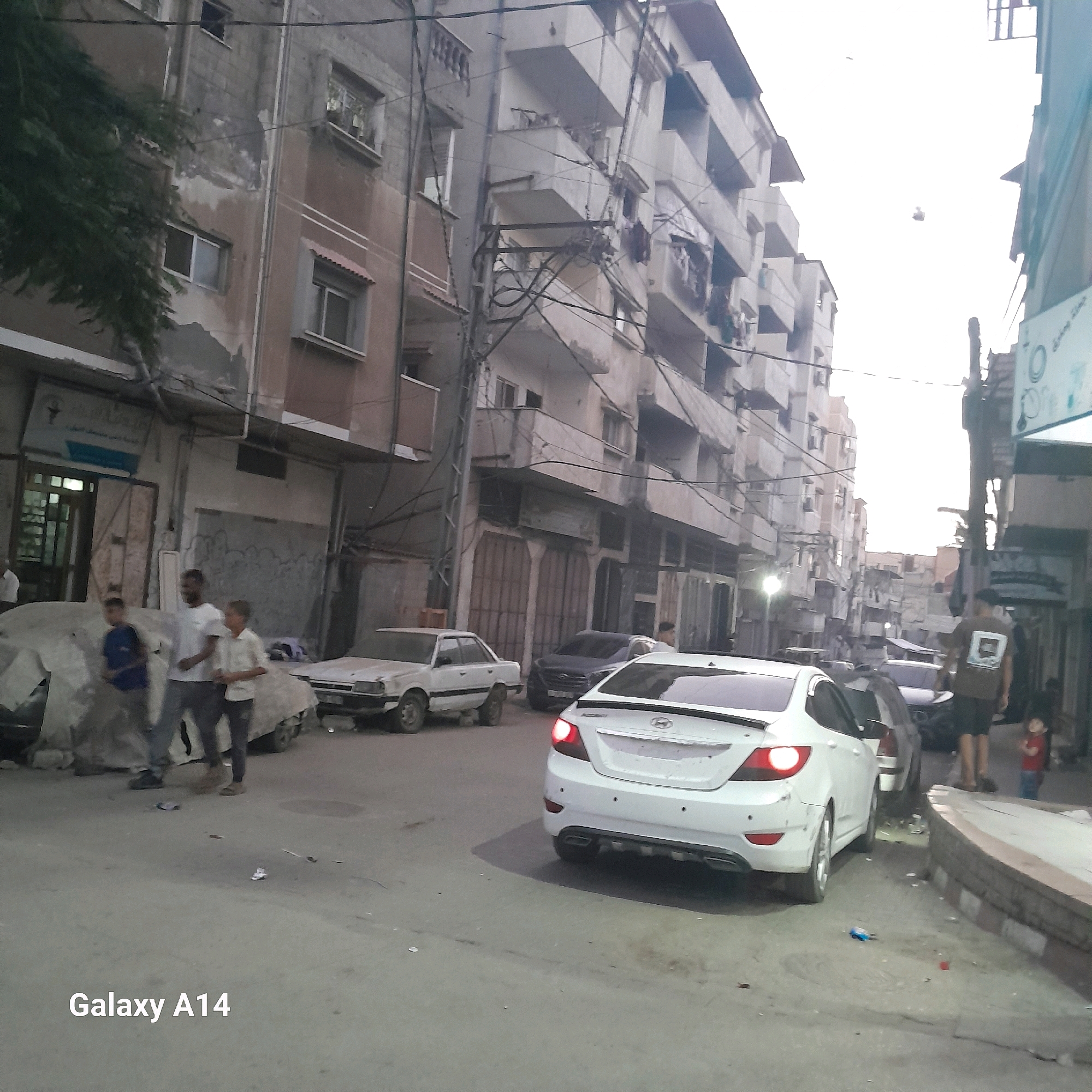 In Deir el-Balah, Palestinian Territories, the image captures a street scene dominated by multi-story buildings. In the foreground, a white sedan is parked. Further back, a dark-colored sedan is also visible. Several people are present on the street. To the left, a person stands near a building entrance. In the middle ground, three individuals are walking, and two cars are parked on the left side of the street, one covered. The buildings exhibit signs of wear, with peeling paint and visible electrical wires strung across the scene. The overall environment suggests a lived-in urban setting.