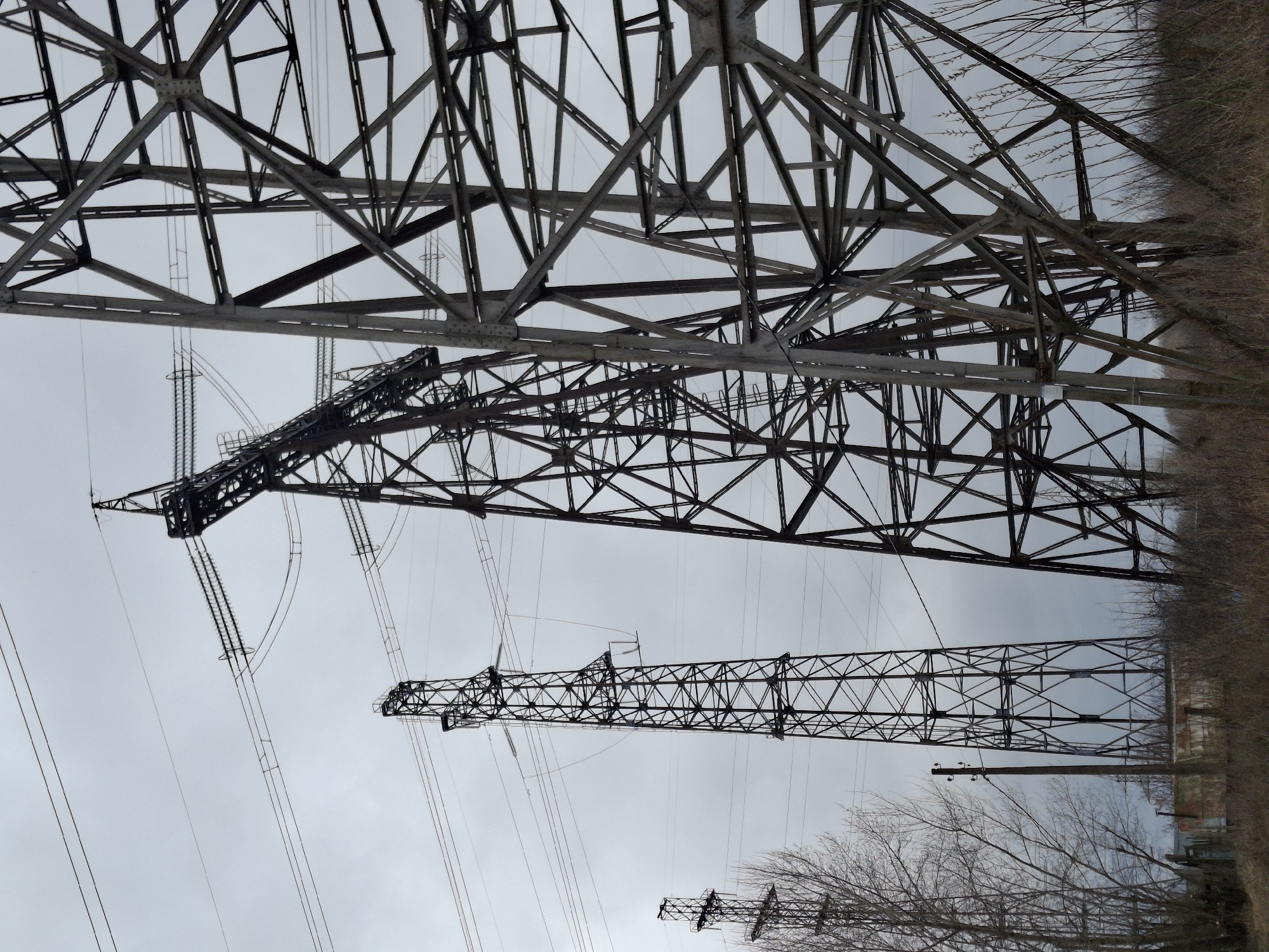 The image shows a view looking up at high-voltage power line towers in what appears to be a rural or industrial area. The sky is overcast. Several towers are visible, constructed from metal lattice. Bare trees and some brush line the perimeter of the scene. The specific location in Chaikovsky, Russia cannot be confirmed solely from the visual data provided. No people are visible in the image. The predominant objects are the power line towers and associated cables.