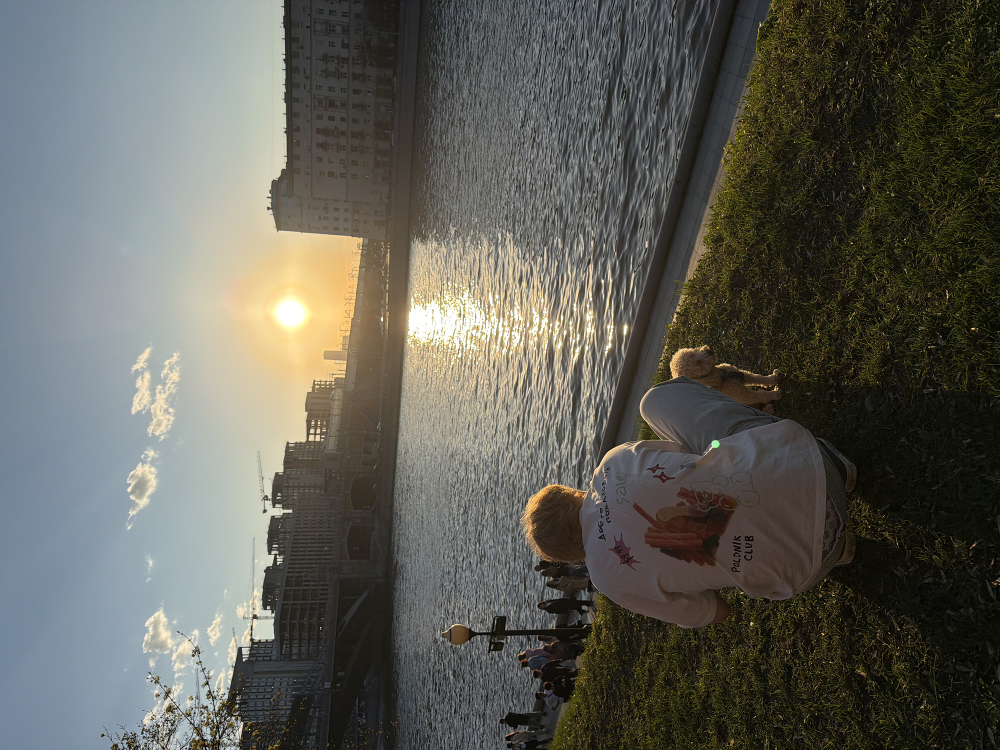The image depicts a river scene at sunset in Moscow, Russia. The sun, low on the horizon, casts a bright glare on the water. Buildings and construction cranes are visible in the background. In the foreground, a person is crouching on a grassy bank beside the river with a small dog. The person is wearing a light-colored t-shirt with designs and text. Other individuals can be seen further down the riverbank. The scene is illuminated by the golden light of the setting sun.