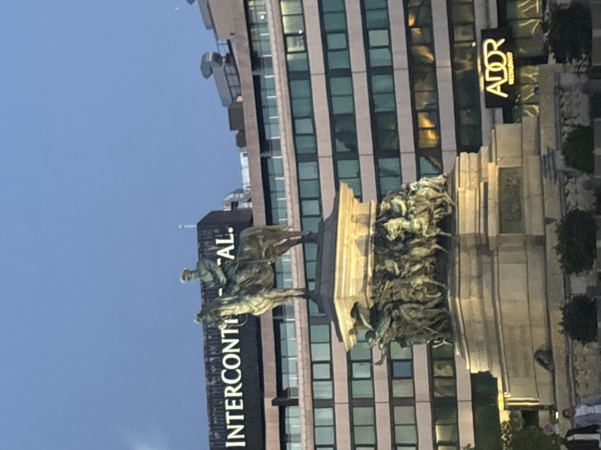 An outdoor, slightly blurry, medium shot shows a bronze equestrian statue in front of a modern building. The statue depicts a man in military attire on horseback, facing left, with intricate relief sculptures at the base of the monument. The building behind the statue is multi-story with a facade of glass windows. A sign on the building reads "INTERCONT..." in large white letters against a dark background, and another illuminated sign below reads "ADOR RESTAURANT" in yellow. The sky is a dusky blue. Two pedestrians are visible in the lower left corner of the frame.