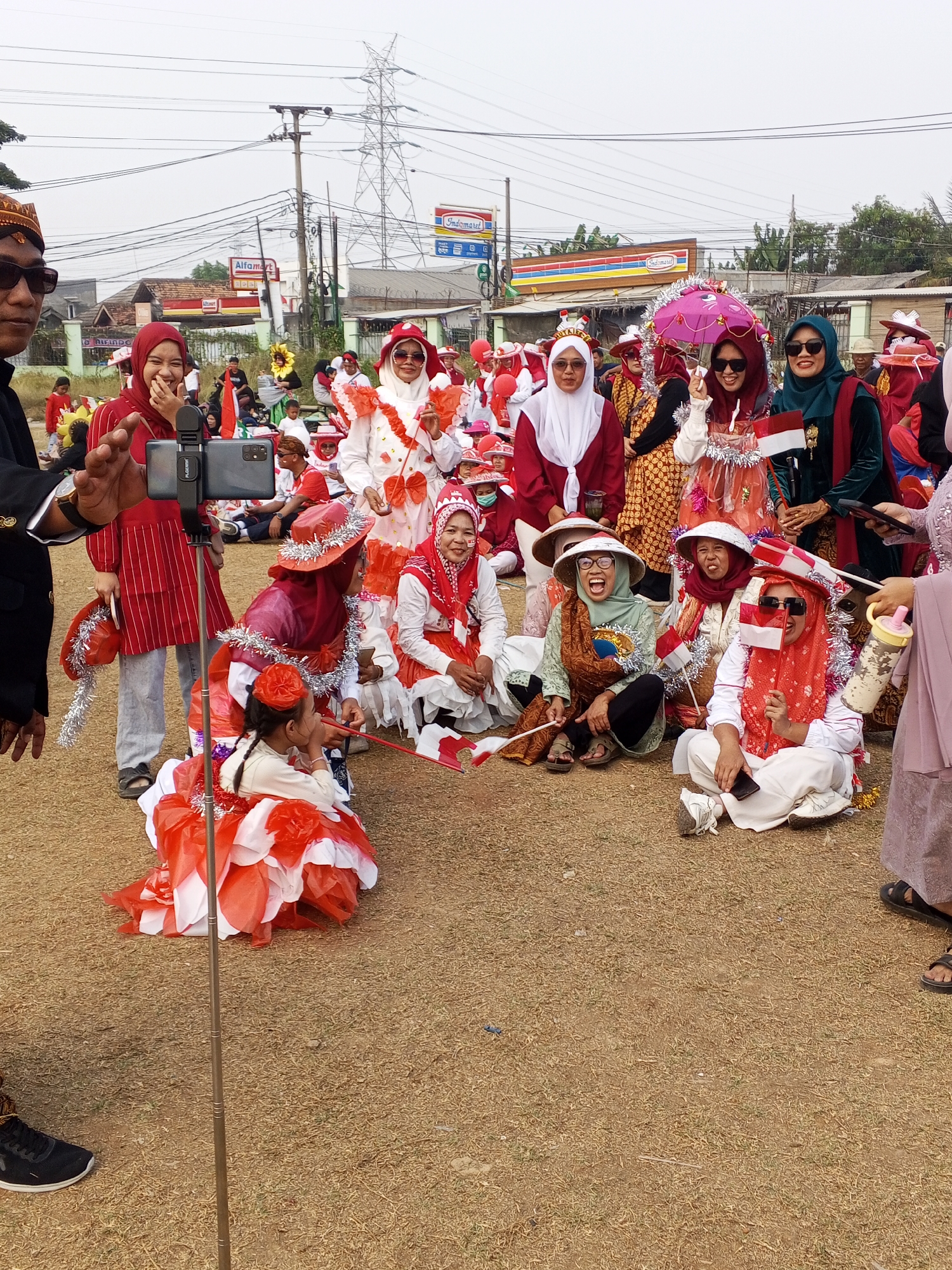 In Ciruas, Indonesia, a group of people, mostly women wearing red and white attire, are gathered outdoors, possibly for a parade or celebration. The scene is characterized by the prominent use of red and white, likely signifying the Indonesian flag. Several individuals are holding small Indonesian flags, and some are wearing festive headwear and decorations. In the foreground, a child in an elaborate red and white costume is seated. A selfie stick with a smartphone is positioned in the center of the frame, suggesting someone is recording the event. The background shows buildings, including what appears to be an Alfamart convenience store, power lines, and utility poles, indicating an urban or semi-urban setting. The ground is covered in dry grass and dirt.