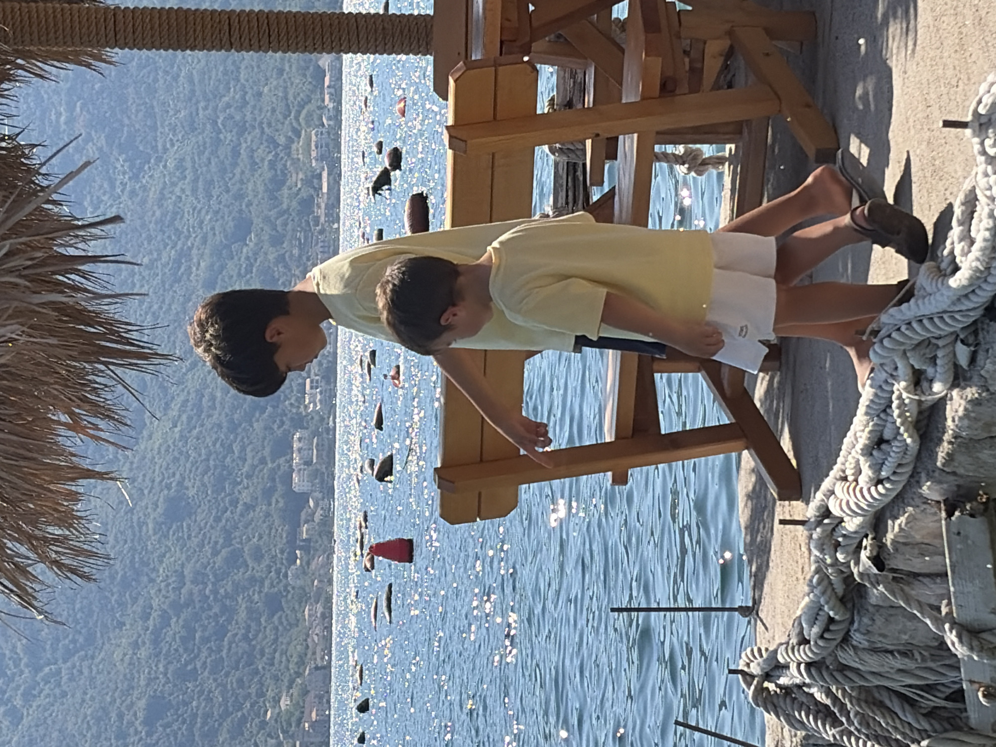 In Dražin Vrt, Montenegro, two young boys in yellow t-shirts are walking along a concrete path next to the sparkling blue water of the Bay of Kotor. The older boy, on the left, is wearing a yellow t-shirt and appears to be pointing towards the water. The younger boy, on the right, is wearing a yellow t-shirt and white shorts. In the background, there are buoys and what appear to be oyster farms in the water, with a lush green, tree-covered mountain range rising behind the bay. A thatched umbrella provides shade over the area, and wooden benches and tables are visible, suggesting an outdoor seating area. Thick ropes are coiled on the stone wall in the foreground. The sun is bright, casting reflections on the water and illuminating the scene.