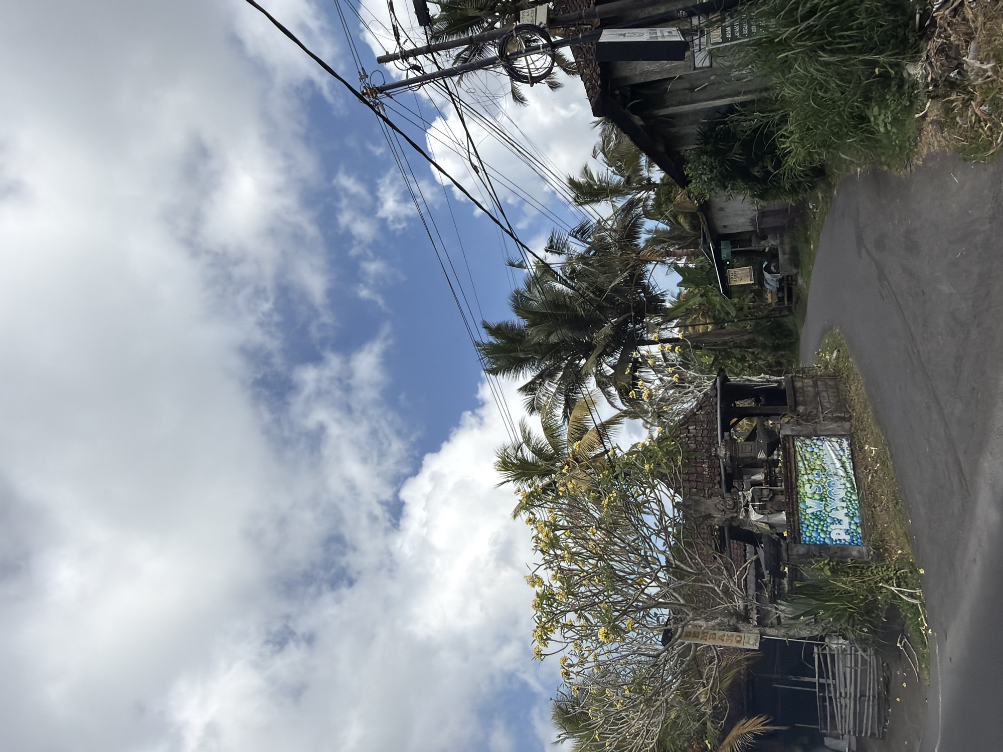 A residential street in Tampaksiring, Indonesia, is depicted under a partly cloudy sky. The road curves to the right, lined with vegetation, palm trees, and low-rise buildings. On the left, a building with a weathered facade and a tiled roof is adorned with a colorful banner that reads "V.S.L PLAYGROUP." A sign with text is visible on a pole in front of this building. To the right, another building with a similar rustic aesthetic features a sign with Indonesian text. Utility poles with numerous wires are prominent in the background, stretching across the scene. The overall impression is that of a tropical, rural setting within a town.