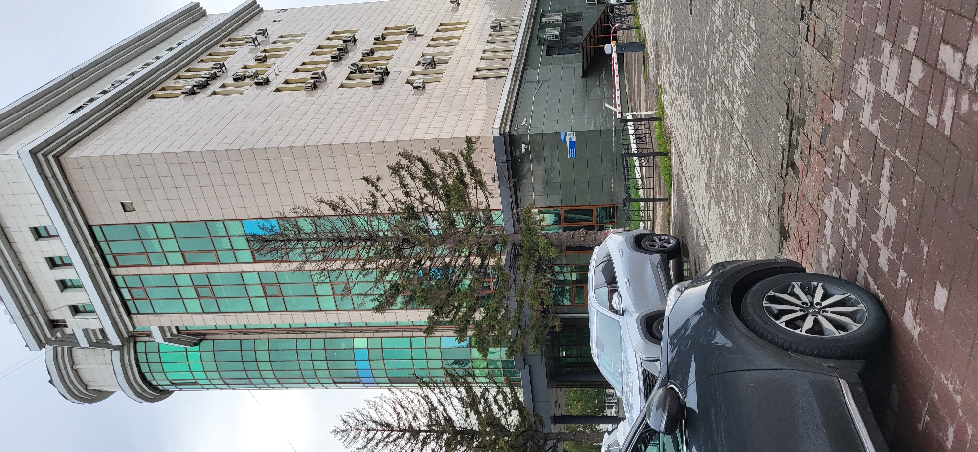 The image depicts a street view in Oskemen, Kazakhstan, featuring a modern office building with a distinctive curved section clad in green glass. The main facade of the building is a light-colored tiled structure with rows of windows and visible air conditioning units. In the foreground, two cars are parked on a wet, red brick paved surface. The car on the left is dark gray, and its wheel and part of its body are visible. To the right of the dark gray car, a silver SUV is parked closer to the building. A black metal fence separates the parking area from the building's grounds. Trees are present in front of the building, with one tall evergreen tree prominently featured. The sky is overcast, indicating a cloudy day. A red and white barrier is visible near the entrance to the building's parking area.