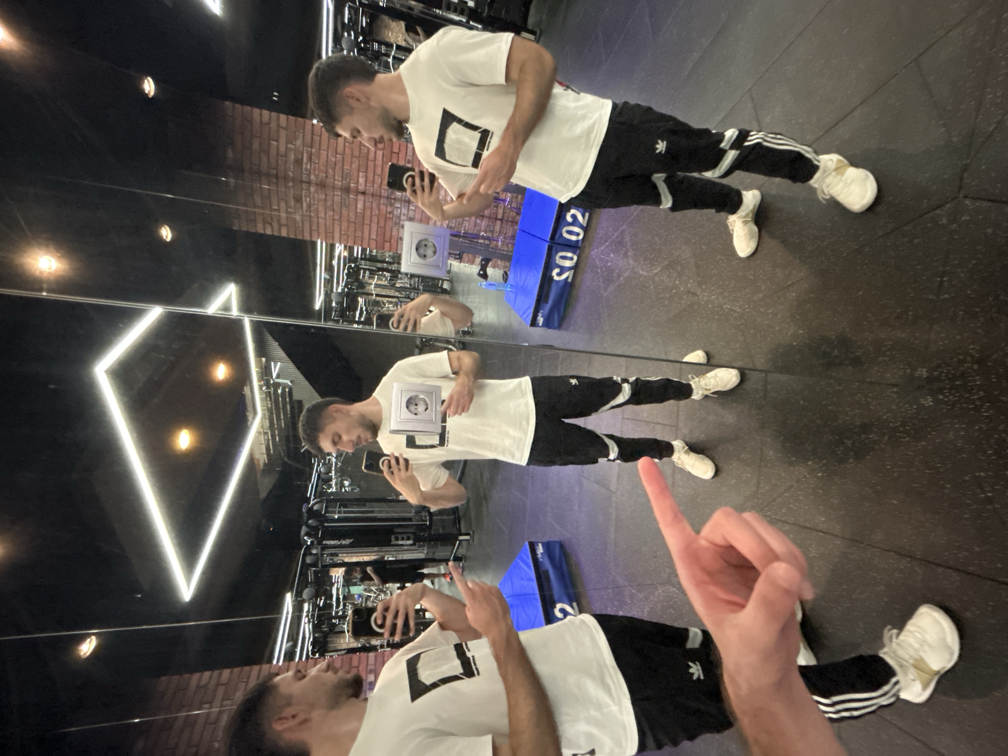 The image shows a man taking a mirror selfie in a gym. The man is wearing a white t-shirt with a black rectangular design, black Adidas track pants, and white sneakers. He is holding a smartphone in his left hand and taking a picture of himself in the mirror. In the reflection, one can see the gym's interior, which includes brick walls, neon lights in the shape of a triangle, and various exercise machines. The mirror reflects the man's image multiple times. One of his hands is raised, pointing a finger towards the top of the frame, partially obscuring the view. Based on the visible gym equipment and interior design, the location is likely a fitness center in Sofia, Bulgaria.