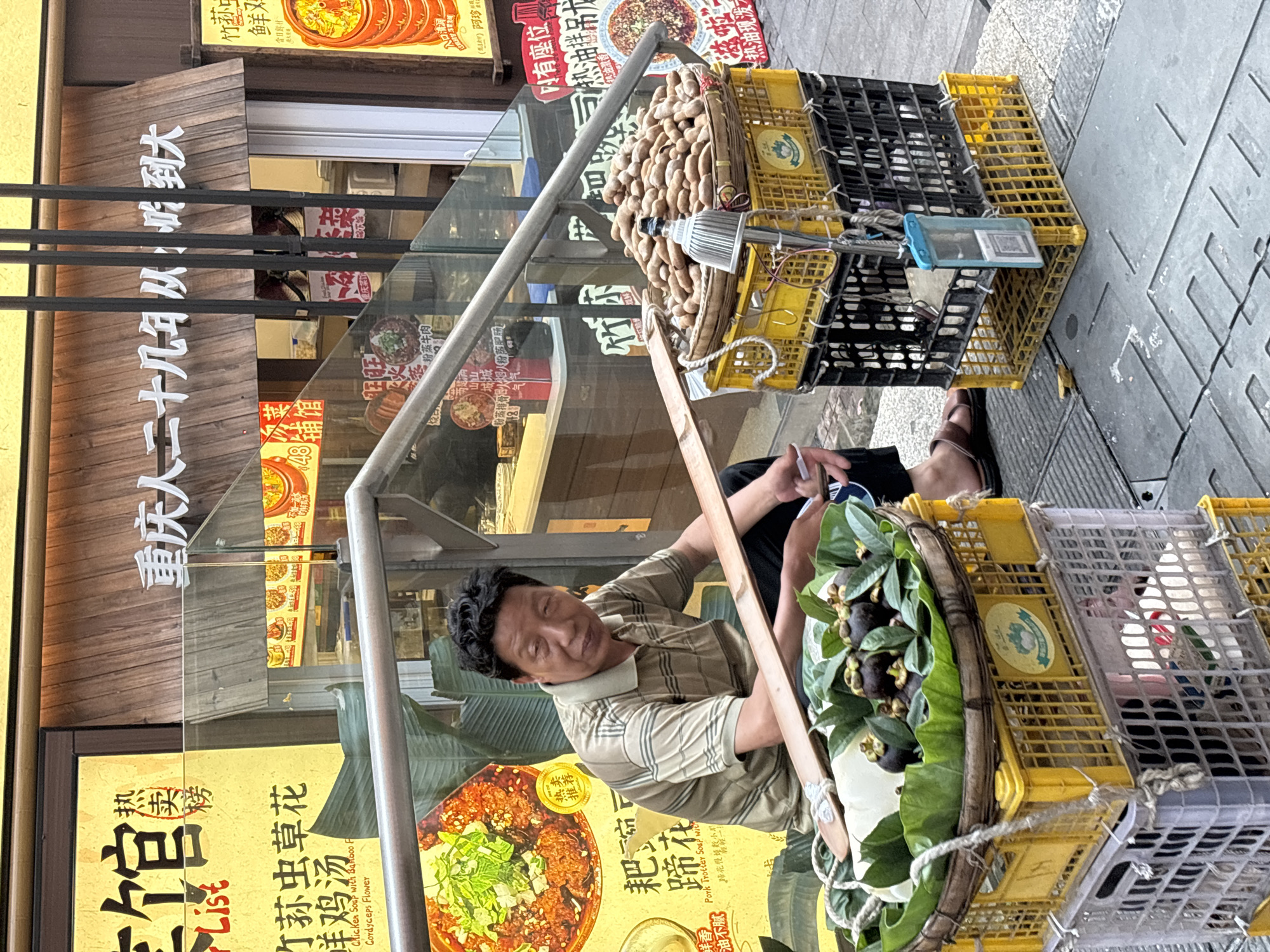 A vendor sits on a sidewalk in front of a restaurant in Yuzhong District, China, selling fruit from a carrying contraption of baskets stacked on top of each other. He is a middle-aged man with short, dark hair, wearing a striped polo shirt and sandals. One basket contains longan fruit, and another contains mangosteen fruit. A metal frame supports the baskets. The restaurant's window displays menus in Chinese.