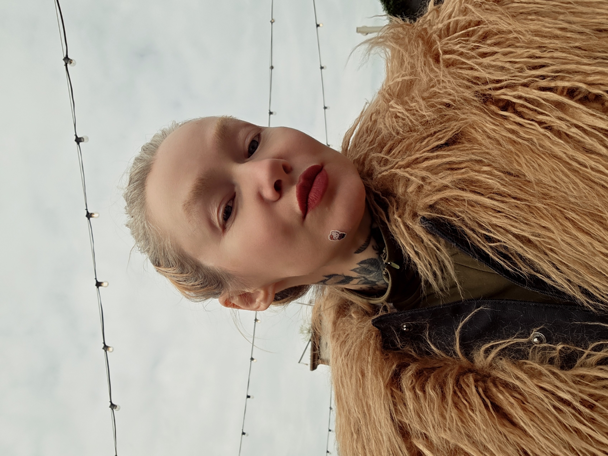 The image presents a low-angle view of a person wearing a shaggy, brown fur coat. The individual has fair skin, light-colored hair styled away from their face, and is wearing dark red lipstick. A small Hello Kitty sticker is visible on their cheek, and they have a prominent tattoo on their neck.
The background shows a sky with string lights hanging overhead, which suggests an outdoor setting. No specific landmarks of Saint Petersburg, Russia are discernible from the image alone.