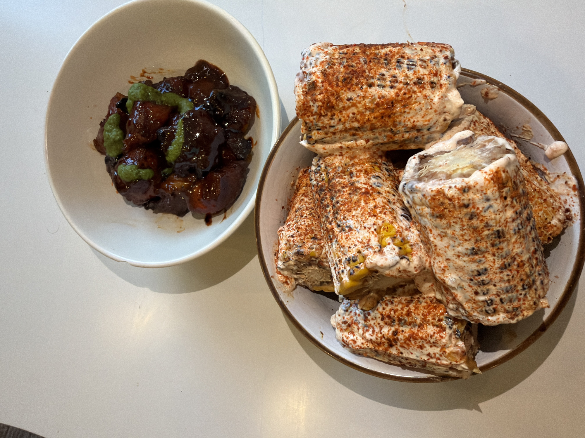 Two bowls of food are presented on a white surface. In the foreground, a round, shallow bowl contains several cobs of corn. The corn is coated in a creamy white sauce, sprinkled with a red spice, and appears to be cut into segments. In the background, a smaller, white bowl holds pieces of dark-colored food coated in a rich, dark sauce and topped with small green dollops, possibly a garnish. The lighting suggests an indoor setting, and no people are visible in the frame. The overall impression is of a prepared meal ready for consumption.