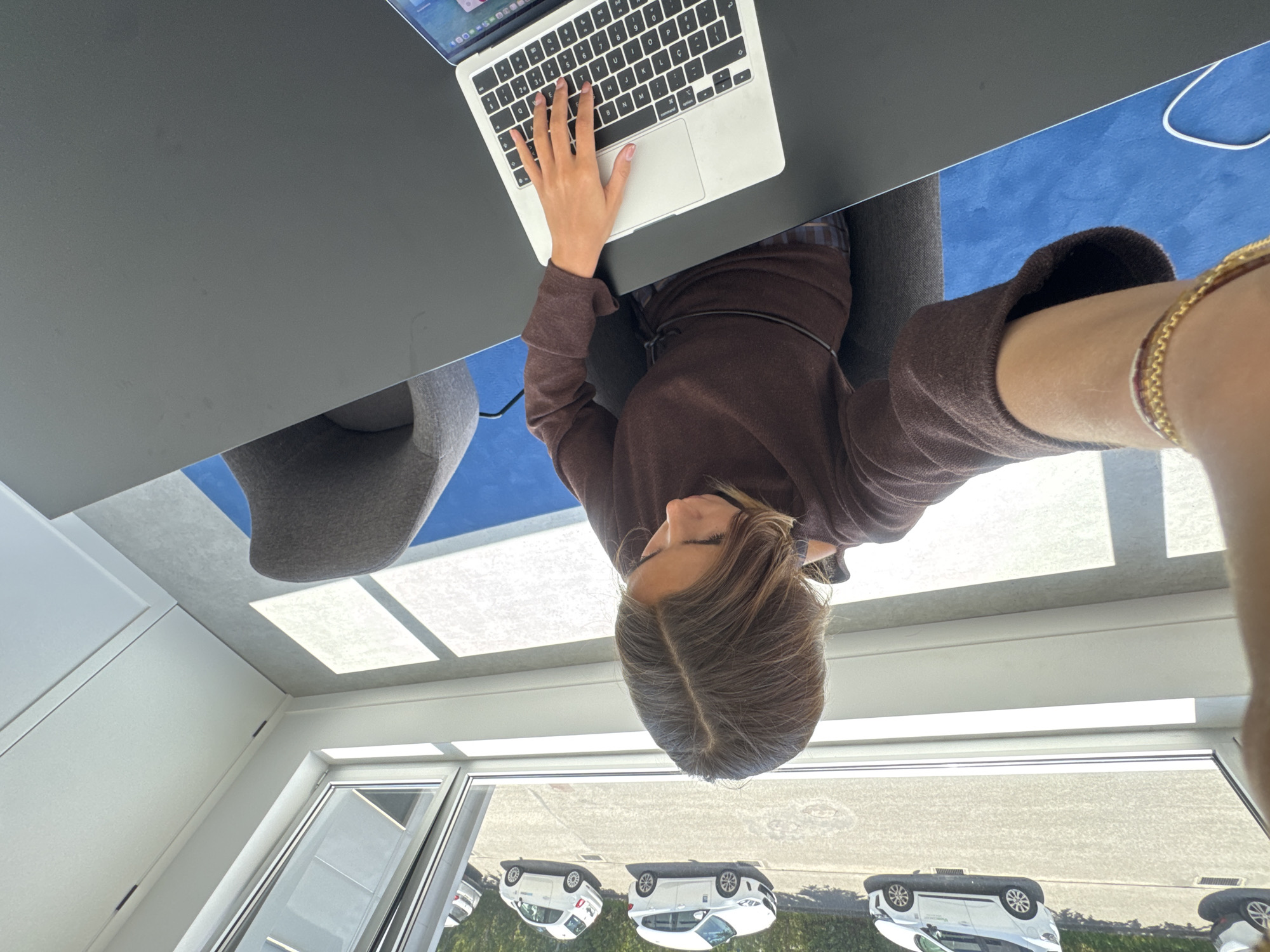 The image, rotated 90 degrees counter-clockwise, captures a young person focused on a laptop in a bright, modern indoor setting, likely an office or showroom, in Torres Vedras, Portugal.
The subject, with light brown hair, is seen from an overhead perspective, wearing a dark brown long-sleeved top. They are seated at a dark, minimalist desk, their right hand actively typing on the QWERTY keyboard of a silver laptop. The laptop screen displays a desktop interface with various application icons. A grey upholstered chair is partially visible under the desk. A gold chain bracelet and a multi-strand bracelet adorn the person's right wrist, which is visible in the foreground, suggesting a selfie from a low angle.
The immediate surroundings include the dark desk surface and, underneath, a section of blue flooring or carpet with a white power cord. The background features a large window or glass wall, through which several white cars are parked outdoors, possibly in a display area or parking lot, bordered by green foliage. The interior ceiling has multiple rectangular light panels, indicating ample natural light during the daytime. The presence of the 'Ç' key on the laptop keyboard aligns with the given location context of Portugal. The scene conveys a sense of focused work or study in a clean, contemporary environment.