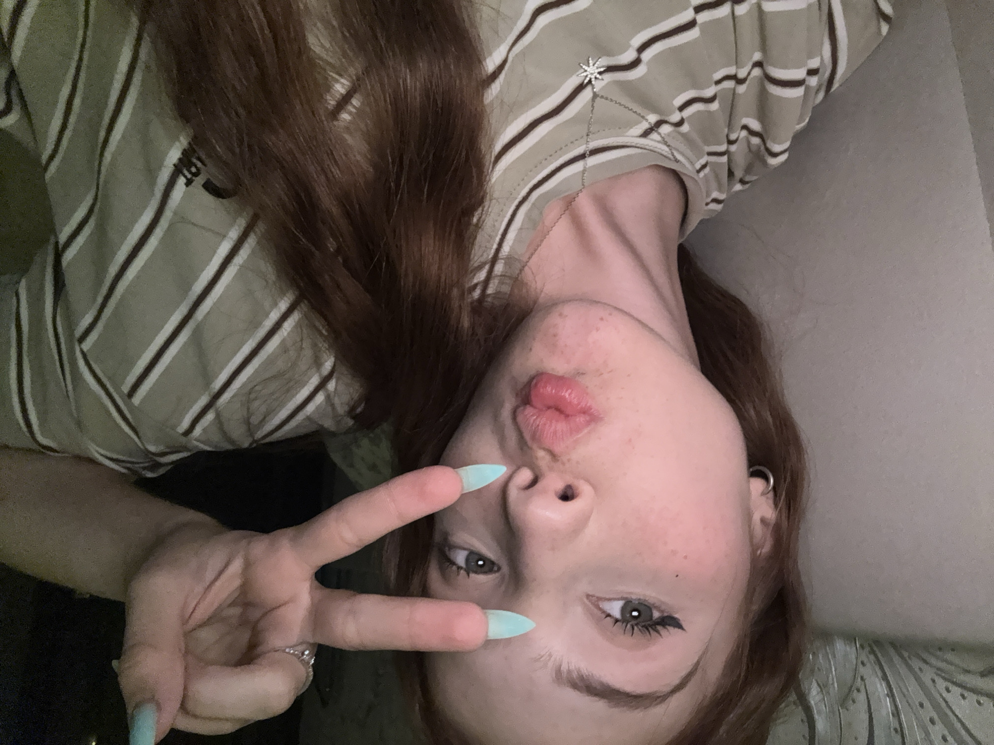 A young woman with long, auburn hair, pale skin, and freckles is pictured. She is wearing a light-colored, striped t-shirt and a silver necklace with a snowflake pendant. Her right hand is raised, forming a peace sign with two fingers extended. Her fingernails are painted a light blue color and appear to be long and sharp. She is also wearing a ring on her ring finger. Her lips are pursed, and her eyes are looking towards the camera. There is a dark background behind her, with some light visible. The image appears to have been taken indoors, likely in a residential setting, and there is no indication of the location being in Naberezhnye Chelny, Russia.