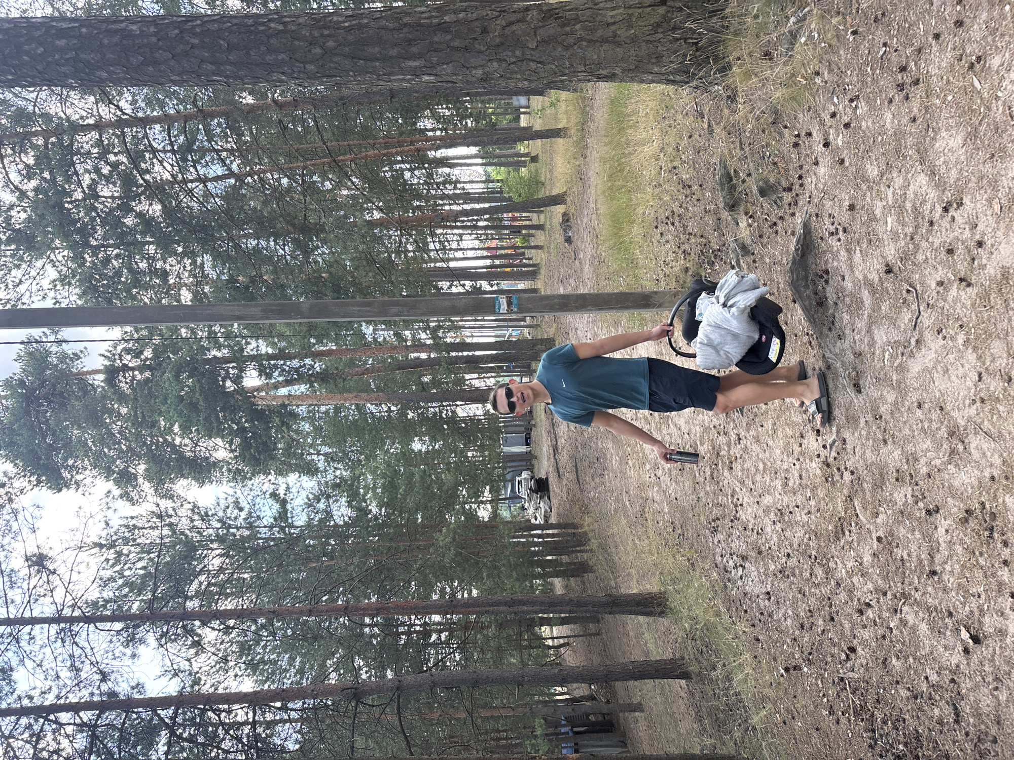 A young man with sunglasses and a green t-shirt is standing on a sandy path in a pine forest. He is holding a can in his right hand and a baby carrier with a gray blanket in his left. The path is littered with pine cones. In the background, there are several pine trees, a wooden pole with a sign, and some buildings and vehicles. The sky is overcast. The location is the city of Kurovskoye, Russia.