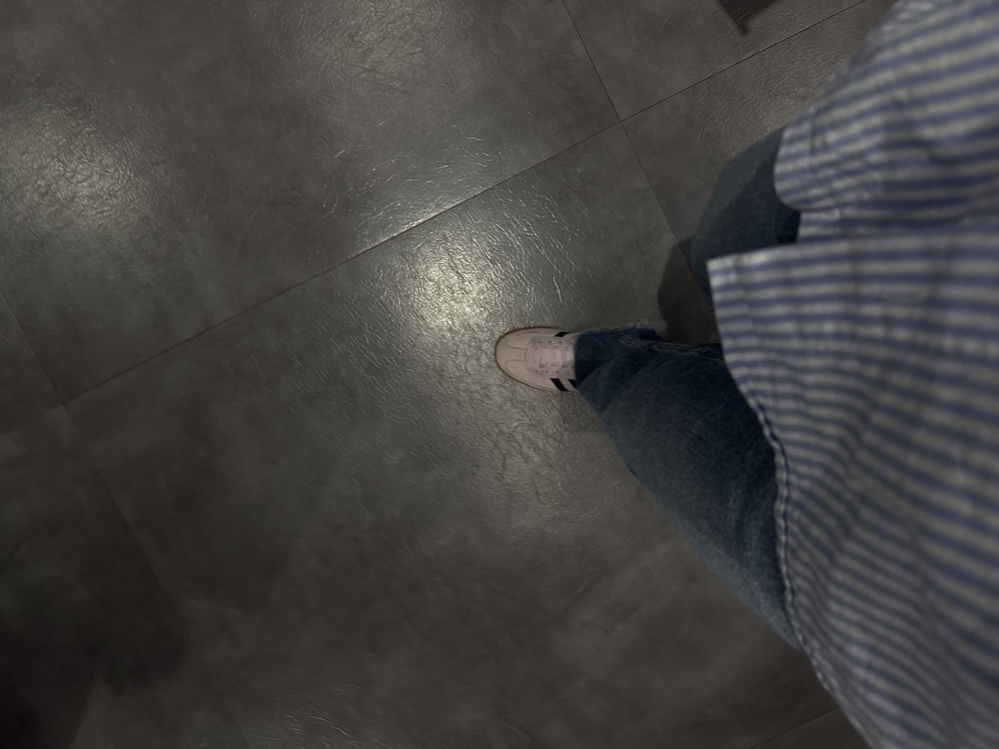 A person wearing blue jeans and a blue and white striped shirt is standing on a dark gray tiled floor. The person is wearing a white sneaker with black stripes.