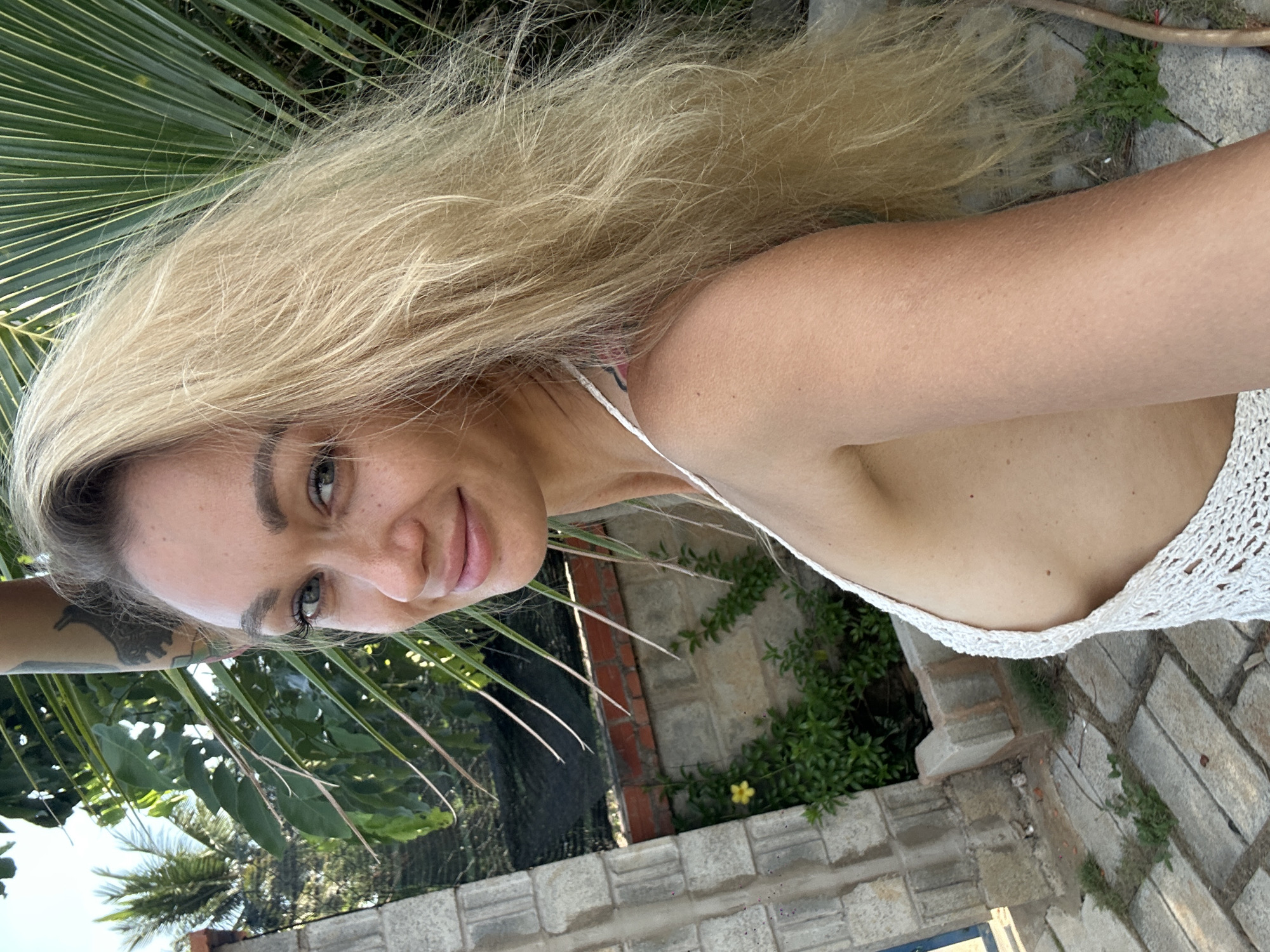 A woman with long, blond hair is pictured from a slightly above, tilted perspective. She has fair skin and is wearing a white, crochet-like top with thin straps. Her arm, which is tattooed, is visible on the left side of the frame. She is smiling slightly and her face is illuminated by natural light.
The background consists of lush greenery, including palm fronds that suggest a tropical location. Part of a brick wall and stone pathway are also visible, indicating an outdoor setting that may be a garden or patio.
The photograph appears to have been taken outdoors, possibly in a backyard. Without more information, it's impossible to confirm if the image was taken specifically in Mũi Né, Vietnam. The environment suggests a warm, tropical climate.