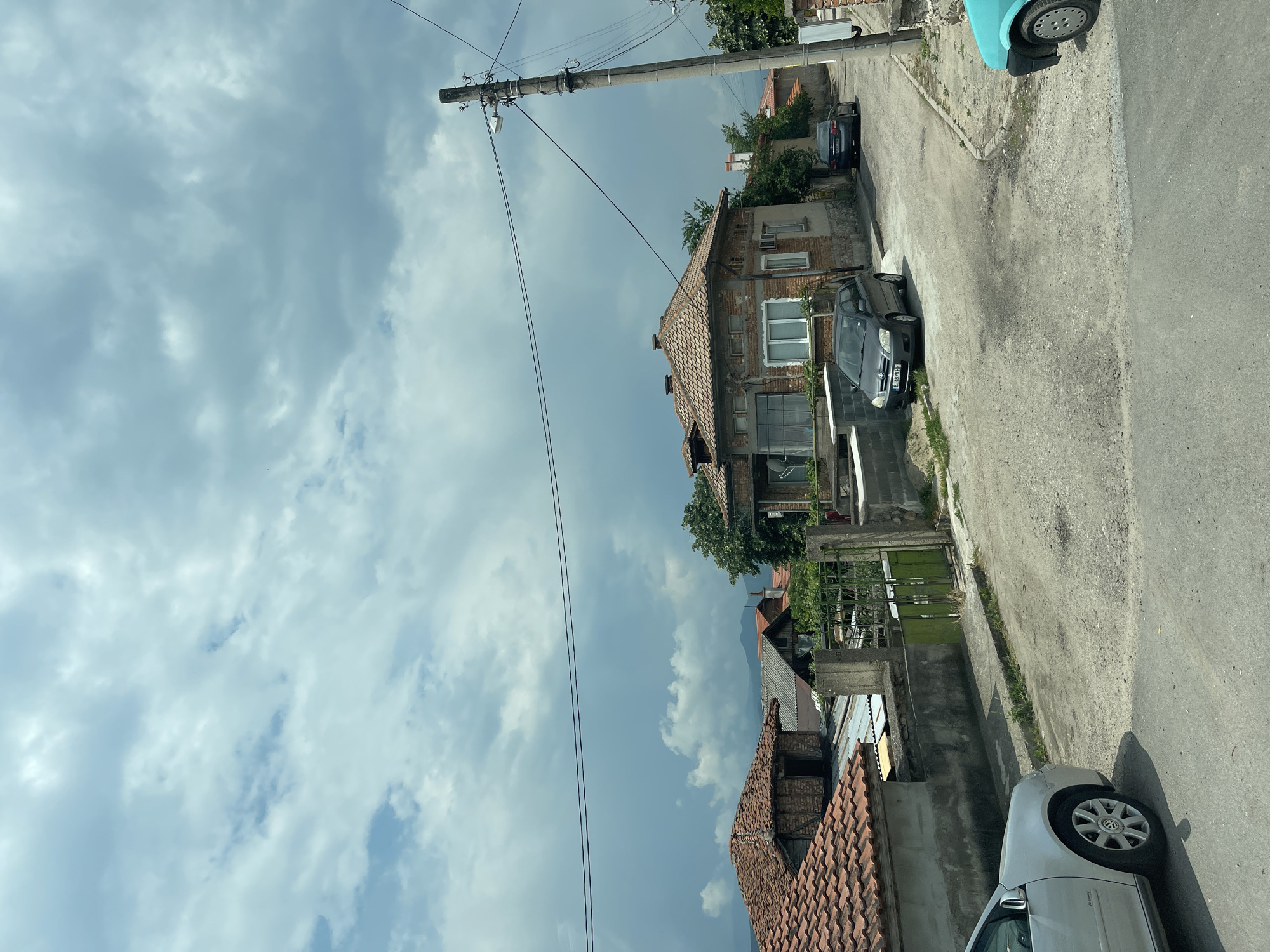 This image captures a static street scene in the town of Karnare, Bulgaria, under an overcast sky filled with dense, light grey clouds.
The street itself is a narrow, unpaved or poorly maintained asphalt road, exhibiting a mix of dirt, gravel, and numerous potholes and uneven surfaces, indicating disrepair. It appears to curve slightly to the right and may have a gentle incline.
On the left side of the foreground, the front-left quarter of a silver Volkswagen Golf is visible. Further down the street on the left, a dark grey Toyota Corolla with Bulgarian license plate B A8761BH is parked beside the road. Adjacent to the road are various residential structures. In the mid-ground left, a house with a reddish-brown terracotta tiled roof is partially seen, and behind it, other buildings with varying roof types, including corrugated metal and older tiles, are present. A weathered green metal gate, flanked by concrete posts, leads into a property.
On the right side of the image, a tall concrete utility pole with multiple cross-arms supporting electrical wires is a dominant feature. A single street light fixture is mounted on the pole. Multiple black wires extend from the pole across the street, creating linear elements in the upper mid-ground. Behind the utility pole and further along the street, a prominent two-story house with a reddish-brown tiled roof and a brick facade, showing signs of wear and aging, features several windows including a large multi-paned window on the lower level. A dark sedan, possibly a Mercedes-Benz, with a partially visible Bulgarian license plate B 4251KM, is parked in front of this house, facing away from the viewer.
In the background, beneath the heavy cloud cover, distant hills or mountains are faintly discernible. Scattered trees and shrubs are visible around the houses, providing limited greenery to the otherwise largely concrete and brick environment. No people are present, and no actions are depicted.
