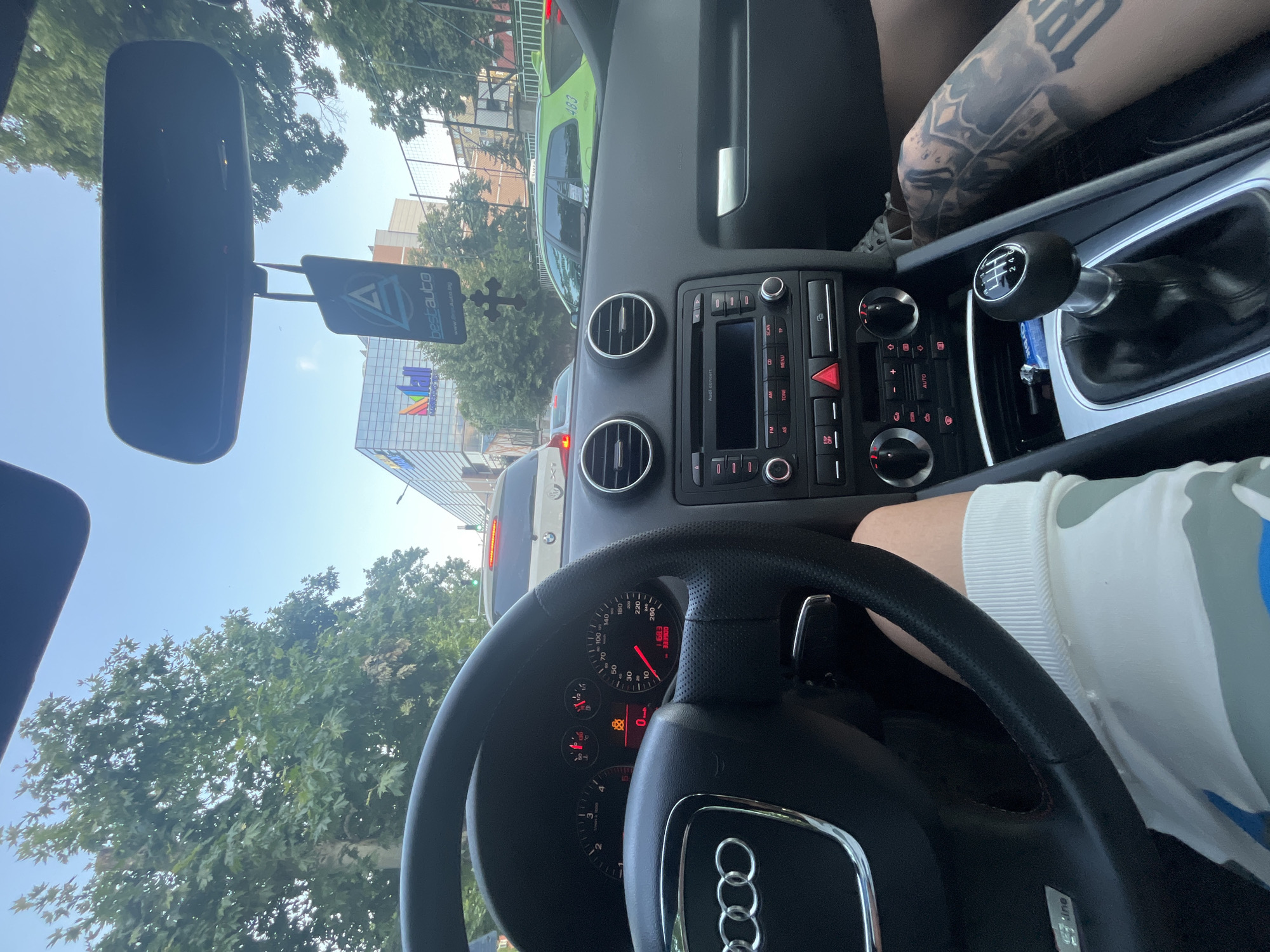 The image captures an interior view from an Audi car, likely an A3 given the dashboard and steering wheel, looking forward through the windshield. The setting is daytime, with clear, sunny weather, and the car appears to be stationary or moving slowly in traffic.
Inside the car, a black leather steering wheel with an Audi logo and a partially visible "S line" badge is prominent in the foreground. The instrument cluster behind it shows the speedometer at 0 km/h, indicating the car is stopped, and an odometer reading of "11603". Several warning lights are illuminated, including a seatbelt warning. To the right, a 6-speed manual gear stick is visible, along with parts of a person's tattooed forearm or upper arm featuring a crown design. Another person's leg, clad in light-colored clothing, is also visible. The center console features an Audi Concert infotainment system with various buttons labeled "MENU", "ECON", "AUTO", and an activated hazard light button.
Outside the car, through the windshield, lush green trees line the street. A green taxi with the number "483" is visible in front, partially obscured by the car's interior. Further down the street, a multi-story building with a distinctive facade featuring colorful, geometric patterns is visible, along with some text that appears to be "МН" or similar. An air freshener or ornament hangs from the rearview mirror, displaying the text "bestauto" and "www.BestAuto.bg", which, combined with the location context, confirms the scene is in Bulgaria, specifically Plovdiv. The sky is bright blue with minimal clouds. The overall scene suggests a typical urban environment.