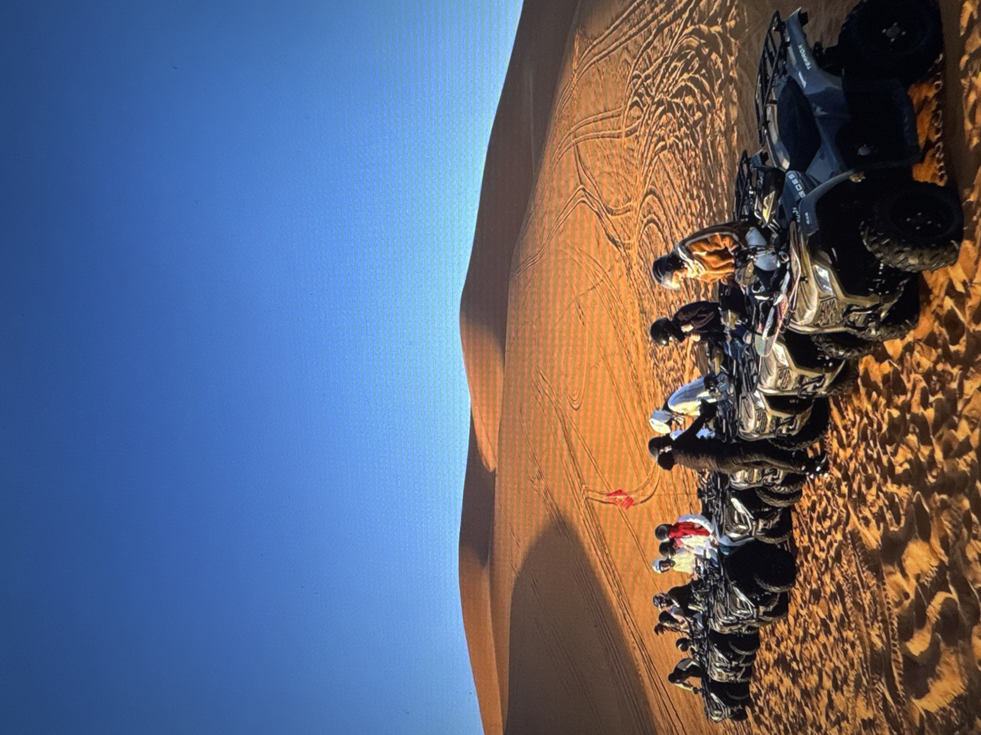 The image shows a group of people wearing helmets and riding quad bikes in a desert landscape. The quad bikes are lined up, and the people appear to be getting ready to ride or have just finished. The desert has large sand dunes and is marked with tire tracks from previous rides. A red flag is visible on one of the dunes. The sky is a clear blue. This scene is not located in Tallinn, Estonia, as Tallinn is a coastal city with a different geographical landscape.